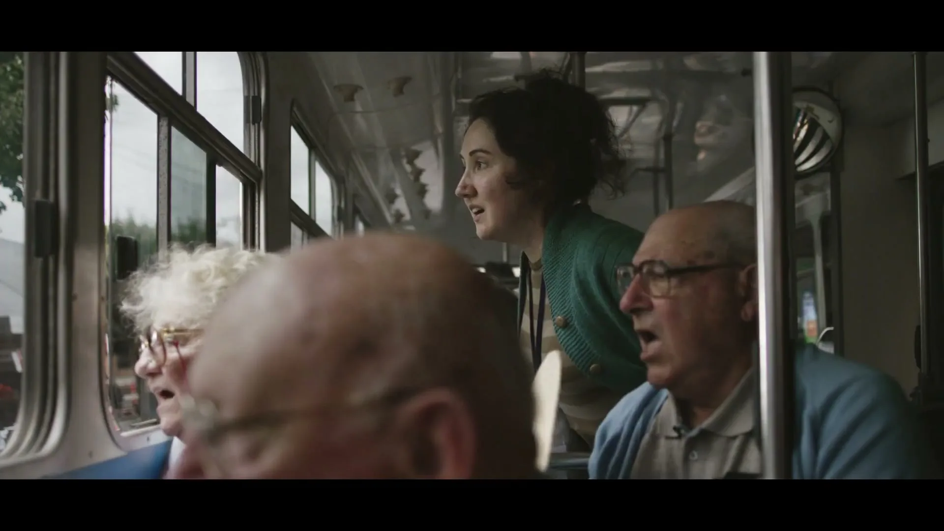 A young woman in a green cardigan and a straw hat looks out the window of a moving bus with two elderly people, all looking surprised.