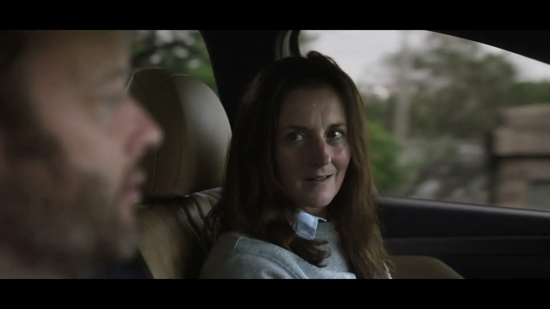 A woman with long brown hair, wearing a grey sweater, sits in the passenger seat of a car, looking at the driver and smiling.