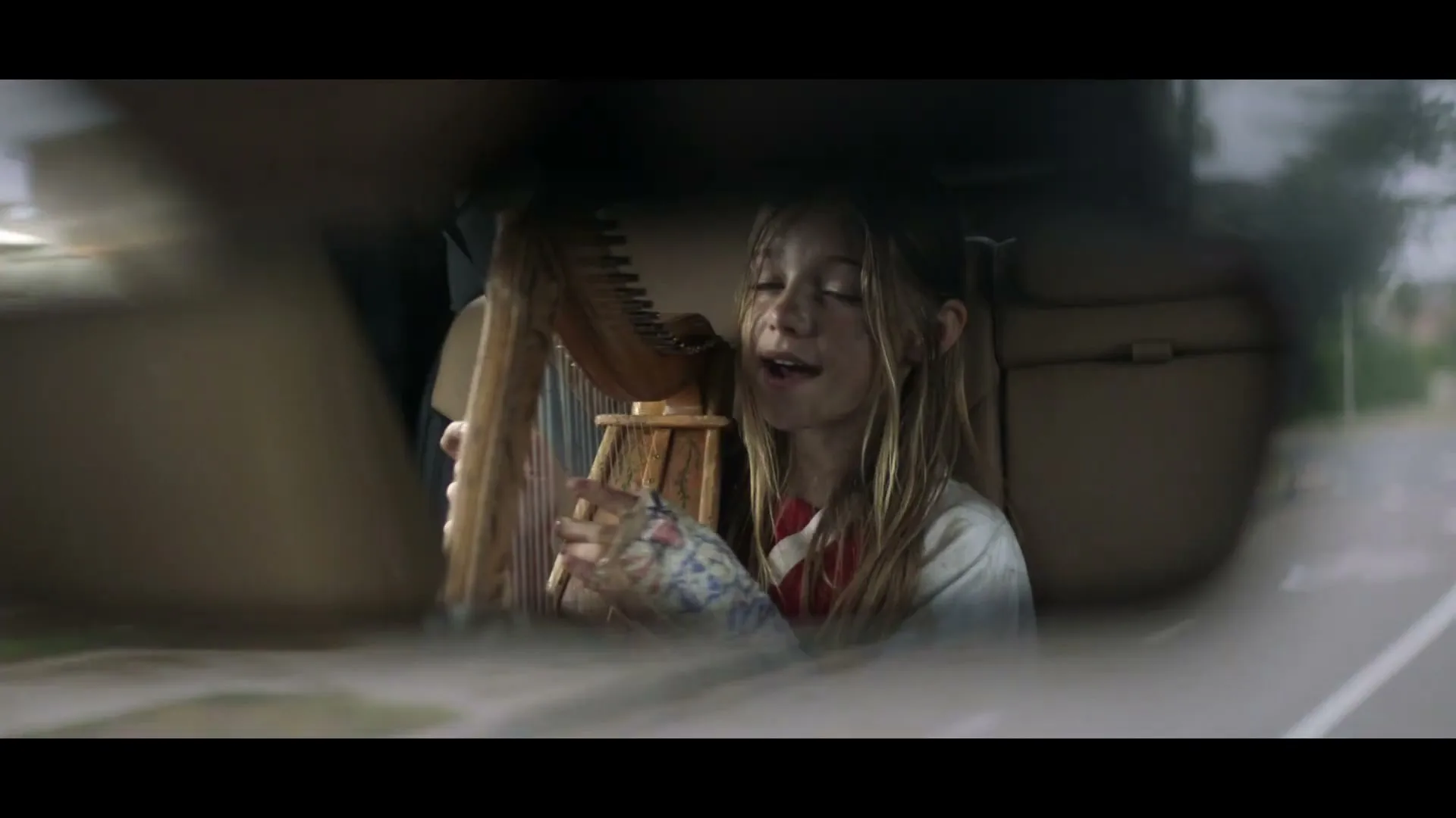A young girl with long blonde hair plays a small harp in the back seat of a car, seen through the rearview mirror.