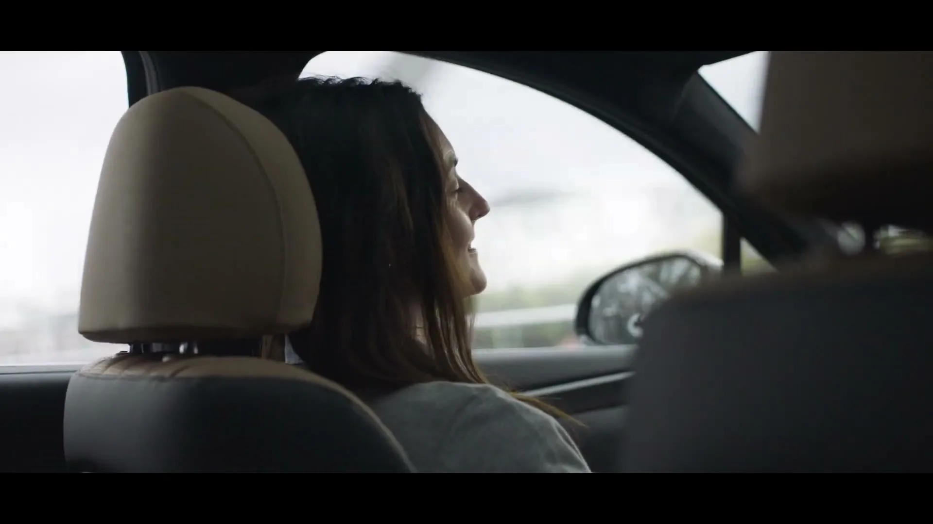 A woman with long brown hair, wearing a light grey shirt, sits in the passenger seat of a car, looking out the window with a slight smile.