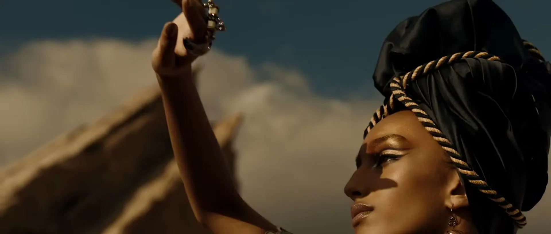 A woman with a black and gold turban and gold makeup raises her hand, wearing a bracelet, against a blue sky and rocky background.
