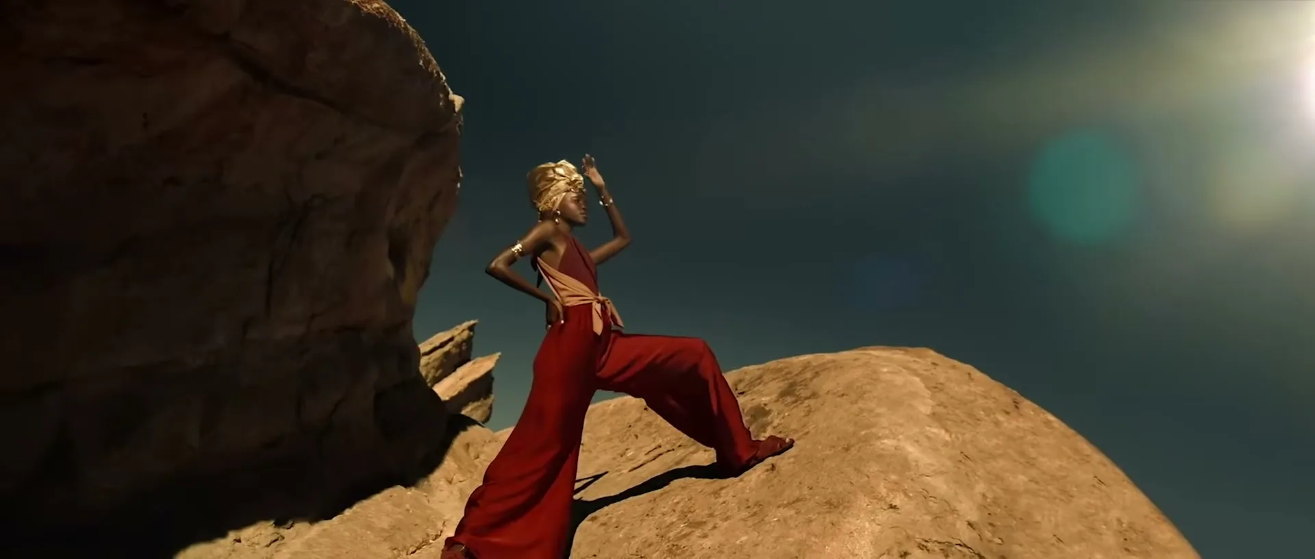 A female model in a red jumpsuit and gold headwrap poses on a rocky outcrop under a bright sun in a desert landscape.
