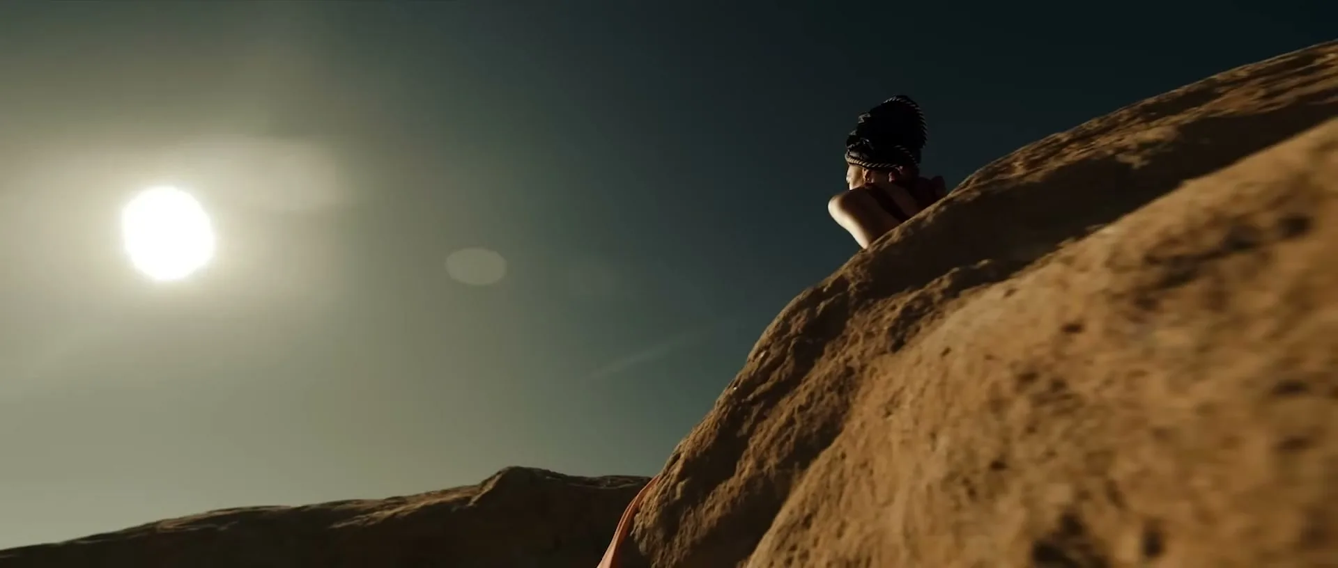 A woman in a black headpiece and top sits on a rocky cliff, looking out at the bright sun in a vast desert landscape.