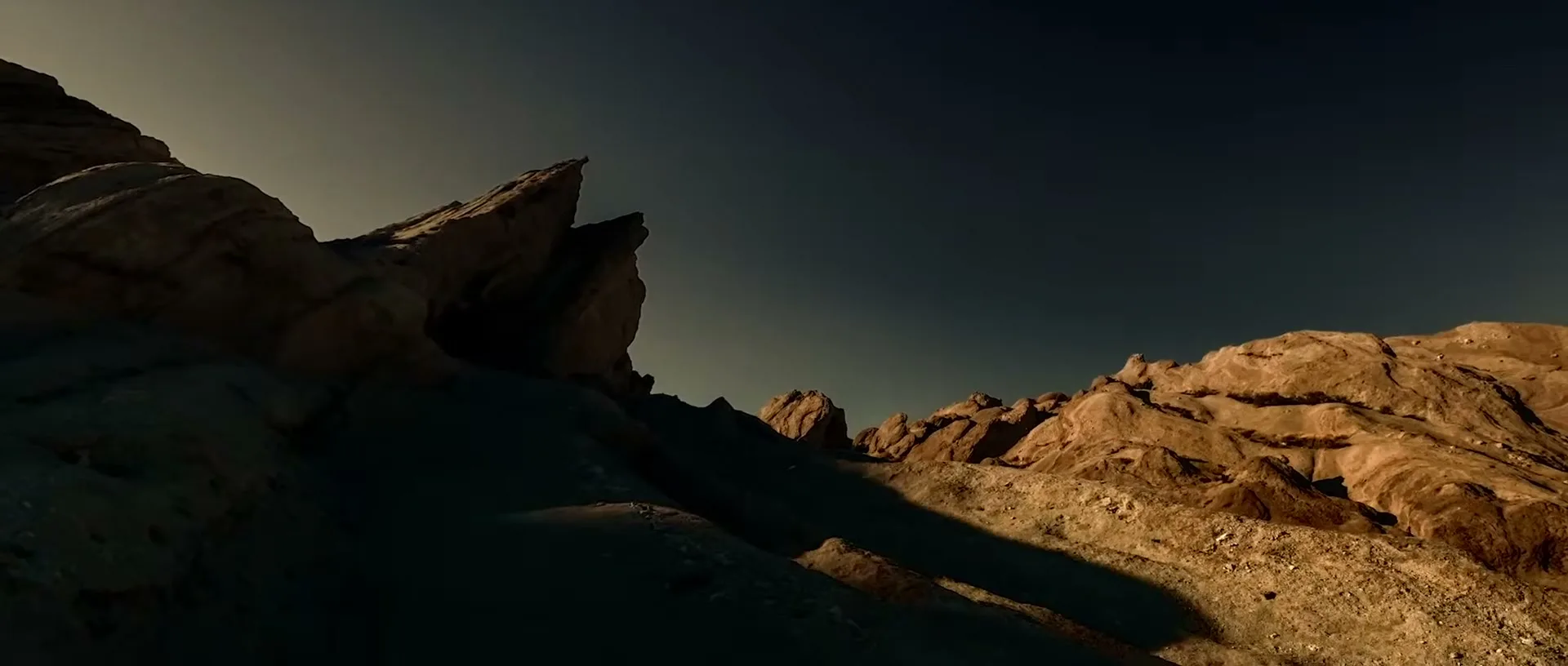 A wide shot of a desert landscape with rugged rock formations under a dark sky, as the sun sets, casting long shadows.