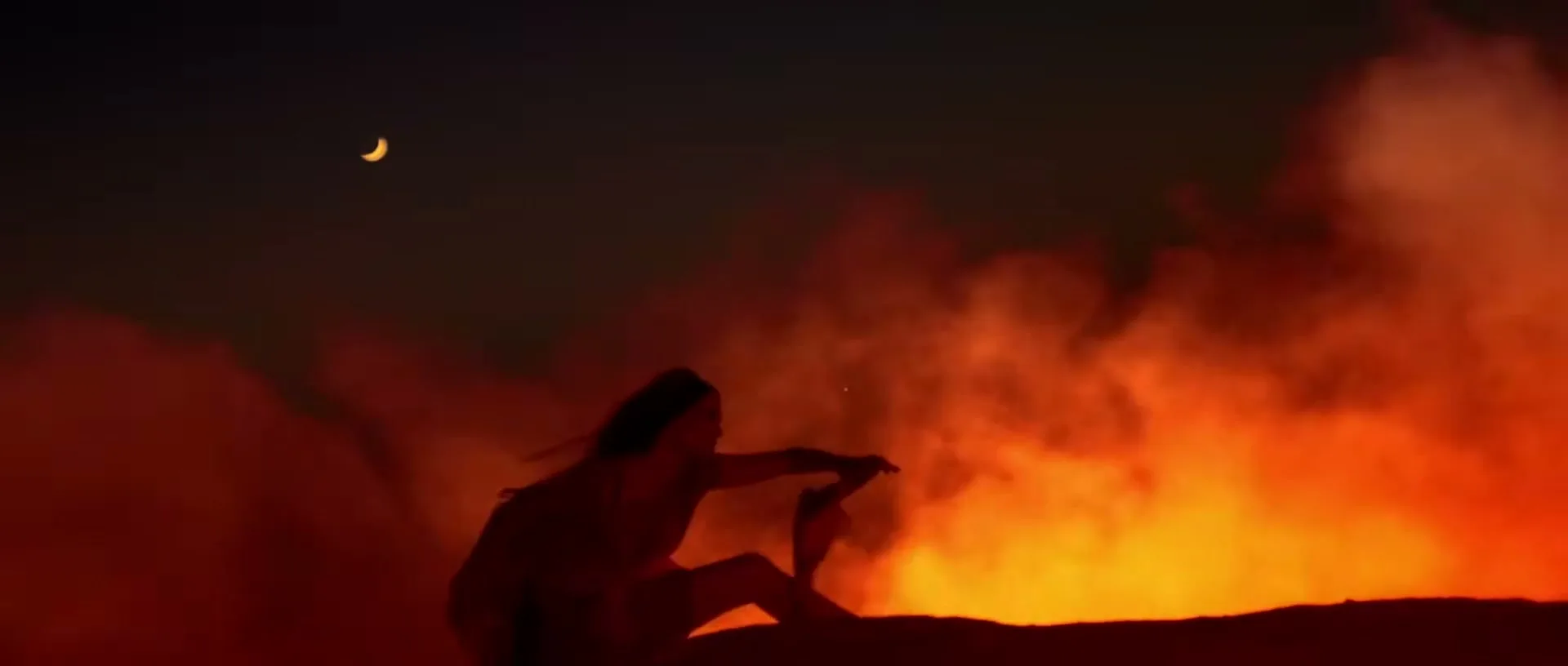 A woman in silhouette sits on a dark ridge against a fiery orange background with smoke and a crescent moon in the dark sky.
