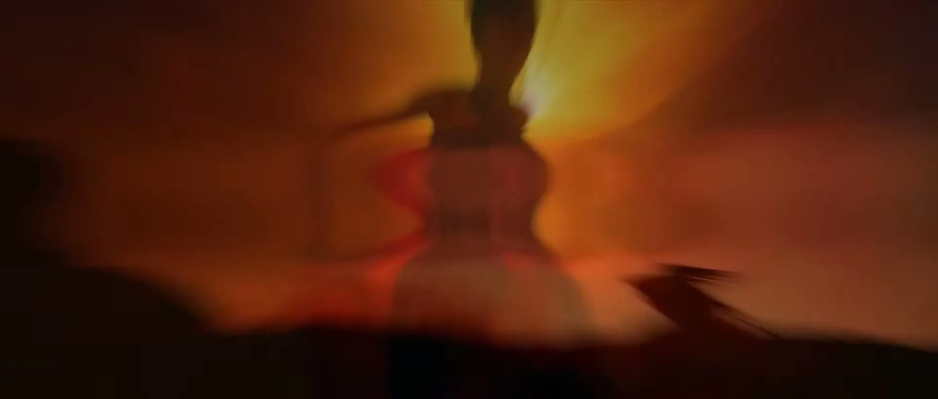 A blurry, silhouetted person stands against a bright, orange-yellow background, with a red object visible on the right.