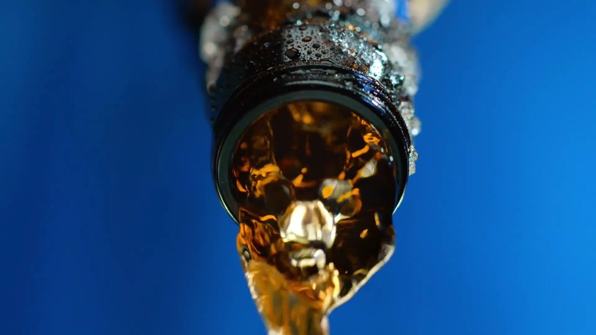 A close-up shot of a cold, wet beer bottle as golden liquid pours out against a vibrant blue background.