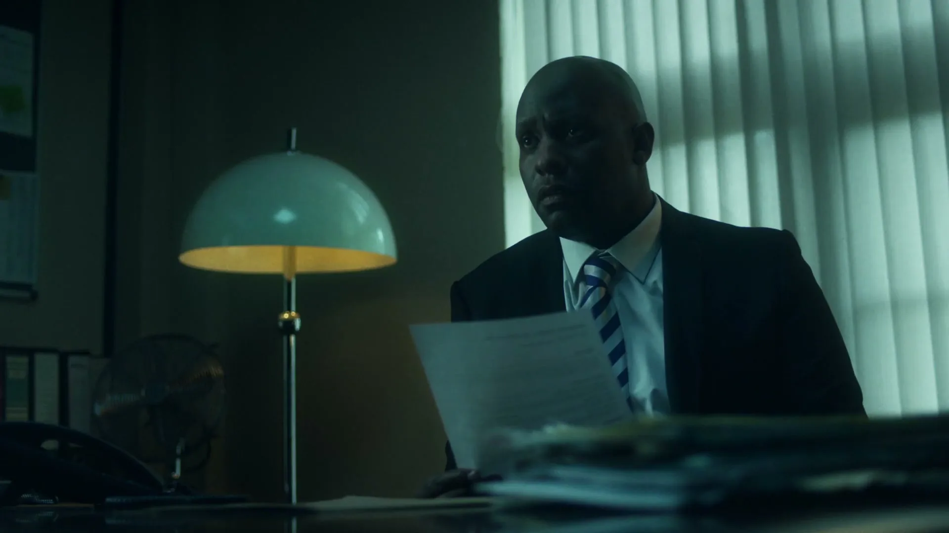 A Black man in his 40s, wearing a suit and tie, sits at a desk in a dimly lit office, looking at papers.