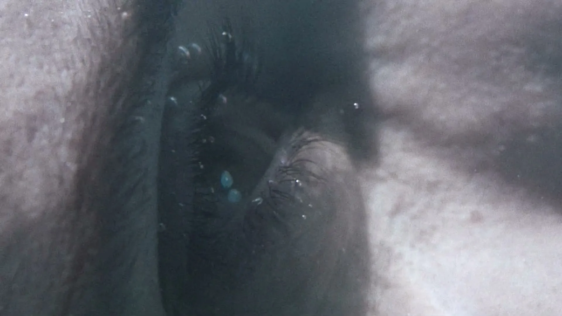 A person's eye is seen in an extreme closeup underwater, with bubbles floating around, creating a blurry and distorted view.