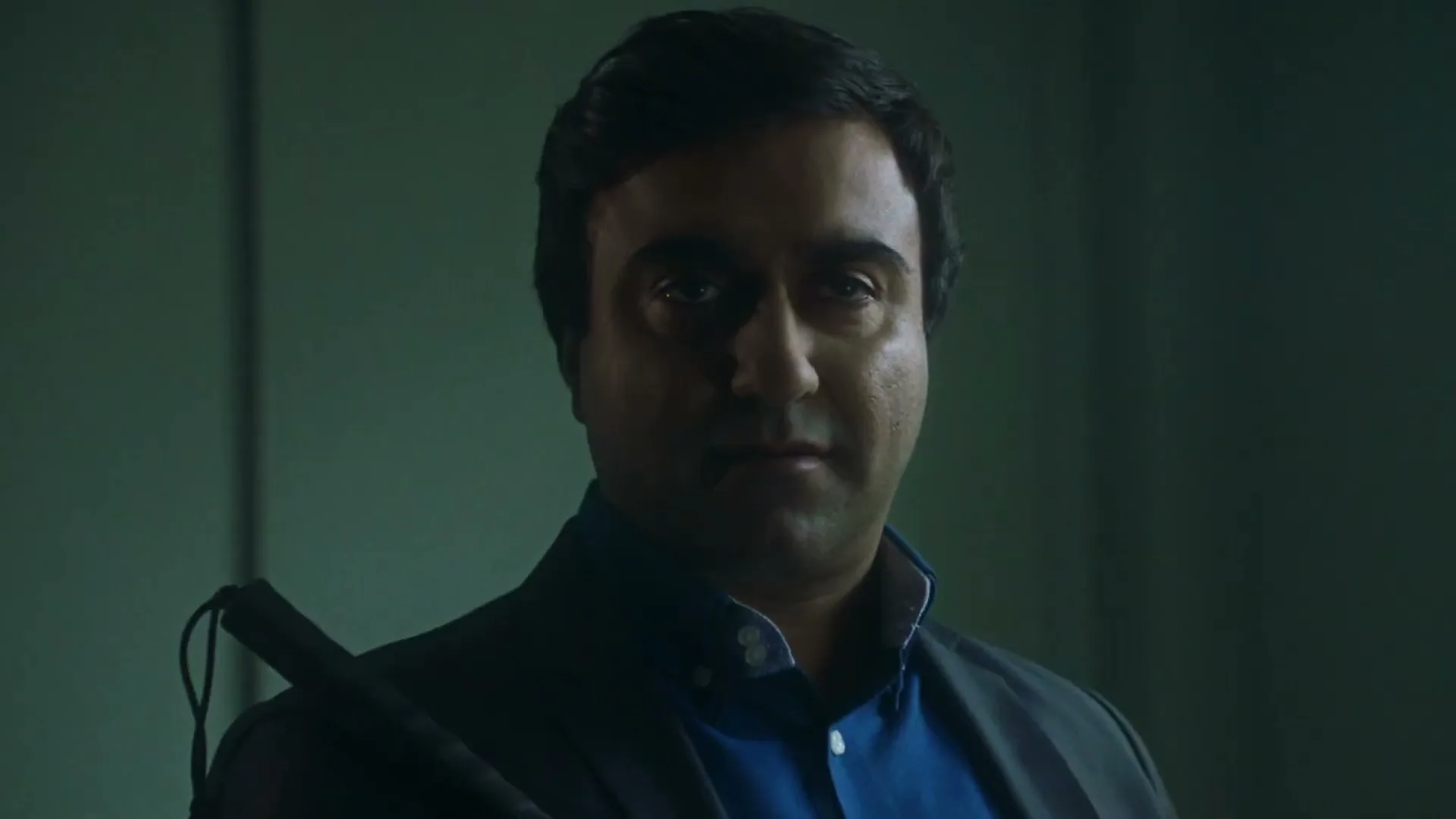 A man with dark hair and a blue shirt holds a white cane, looking directly at the camera in a dimly lit room.