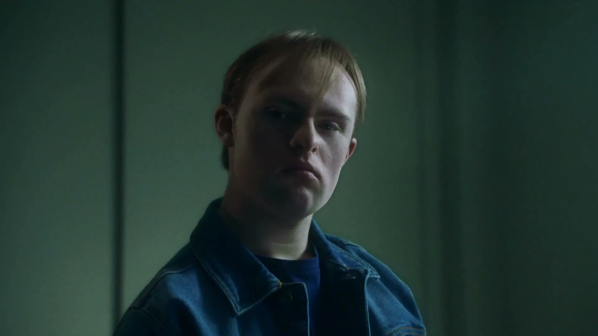 A young man with Down syndrome, wearing a denim jacket, looks directly at the camera with a serious expression in a dimly lit room.