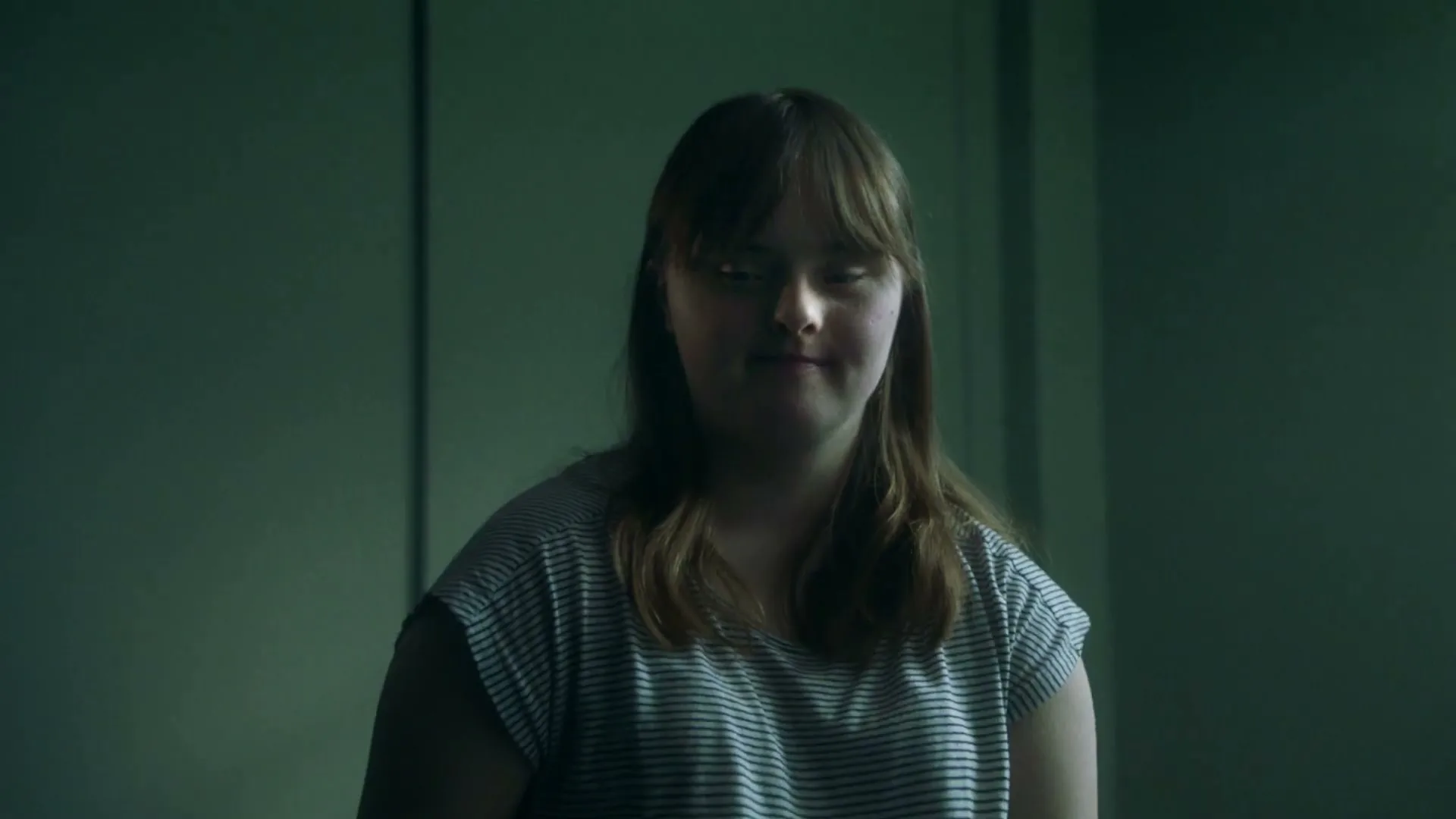 A young woman with Down syndrome, wearing a striped t-shirt, looks directly at the camera with a slight smile in a dimly lit room.