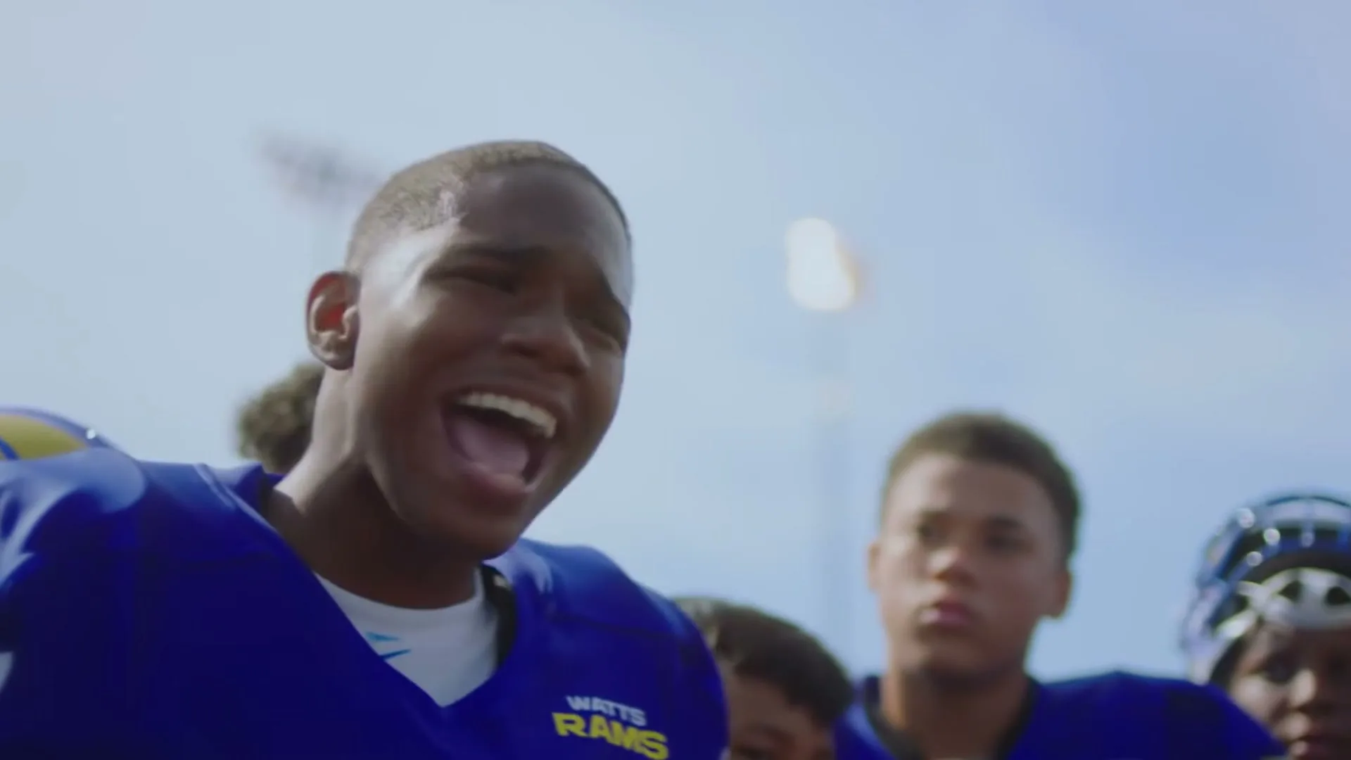 A young male football player in a blue and yellow uniform with "WATTS RAMS" on it, stands with his teammates on a sunny football field, giving a motivational speech.