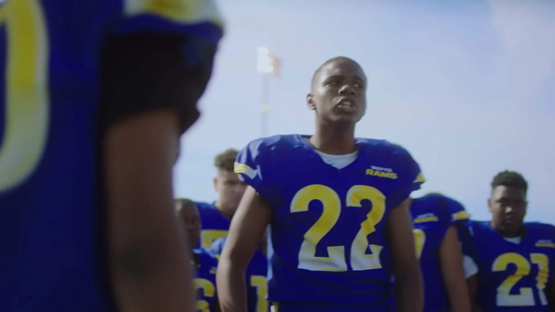 A young male football player in a blue jersey with number 22 on it, stands with his teammates on a sunny field, looking determined.