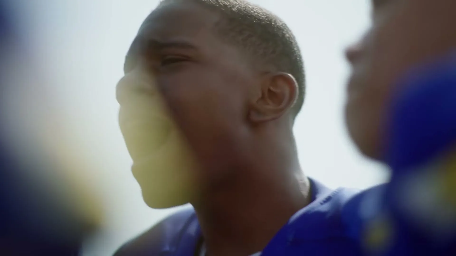 A young Black male football player in a blue jersey shouts with his mouth open, surrounded by teammates on a sunny day.