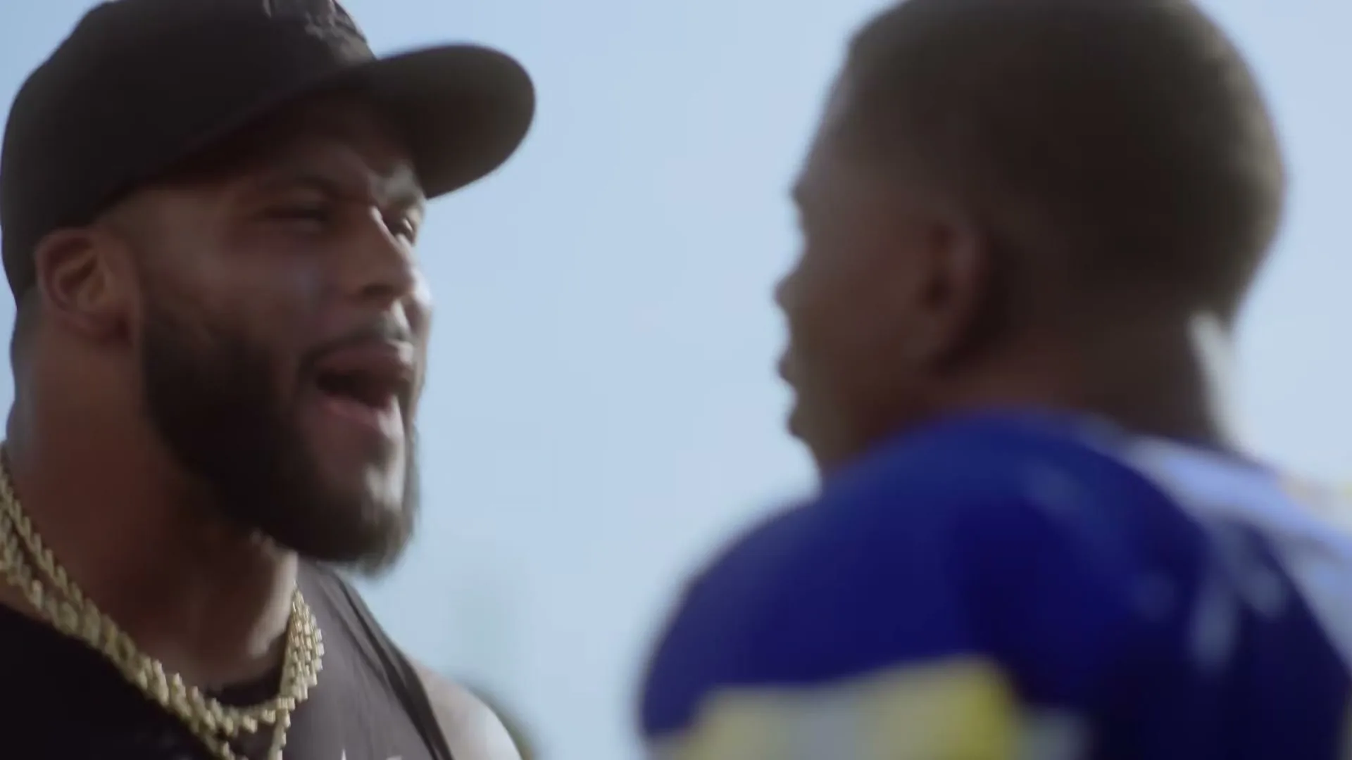 A muscular man in a black tank top and cap, wearing a gold chain, laughs while facing another man in a blue jersey, outdoors under a bright sky.