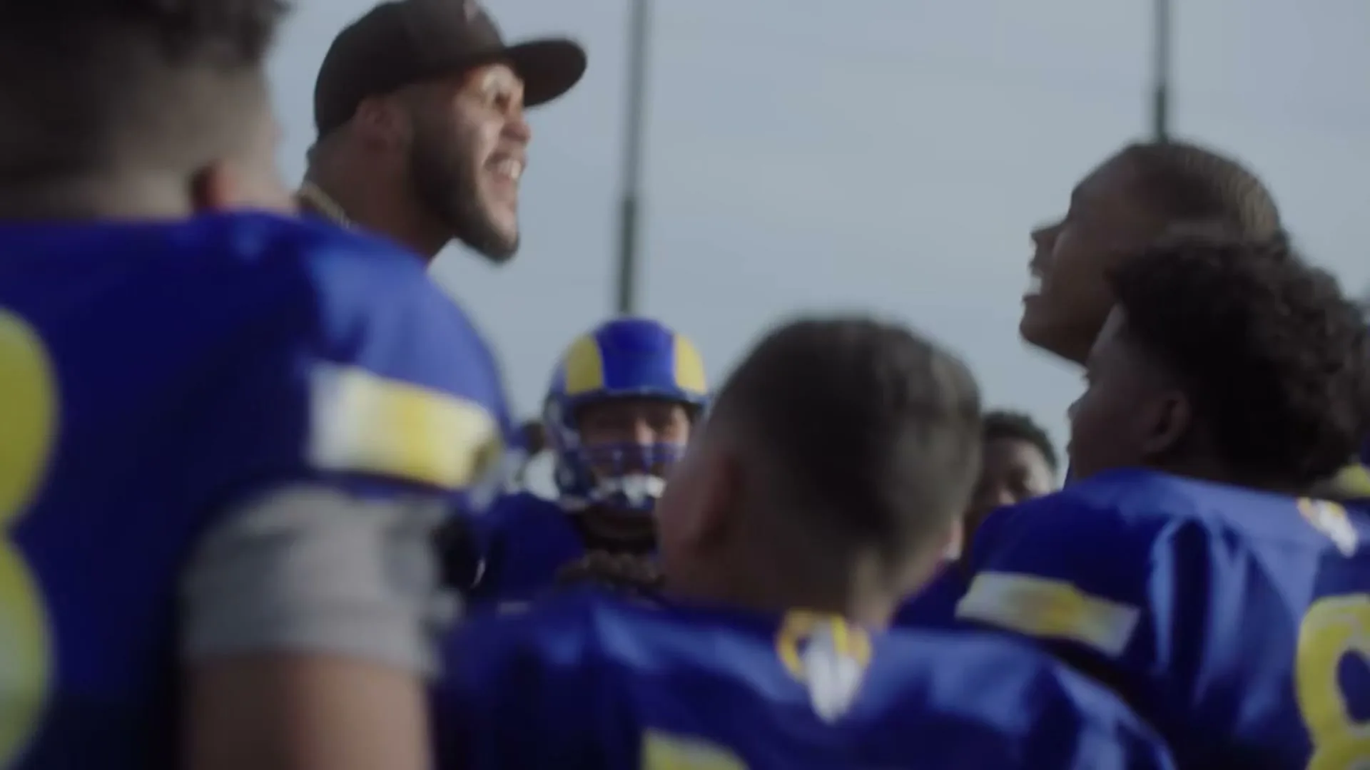A football player in a black cap and tank top, wearing a gold chain, shouts at a group of young football players in blue and yellow jerseys, on a sunny outdoor field.