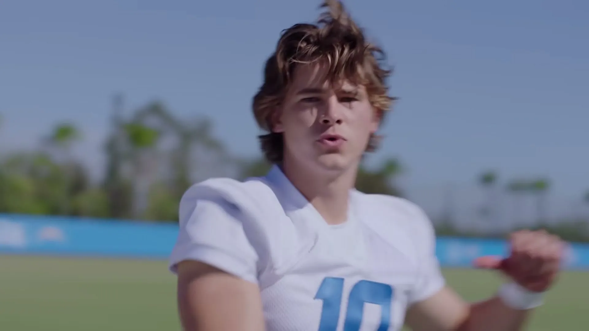 A young male football player in a white jersey throws a football, then turns to the camera and makes a thumbs down gesture on a sunny field.