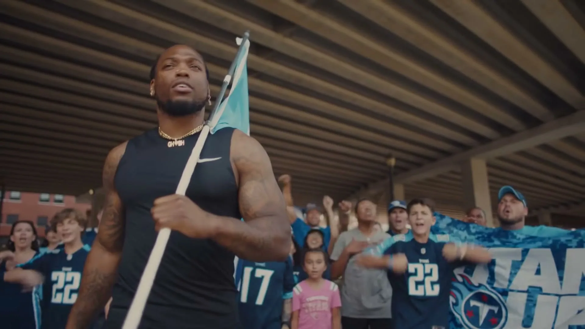 Derrick Henry, a muscular Black man, leads a crowd of cheering fans, holding a light blue flag under a concrete overpass.
