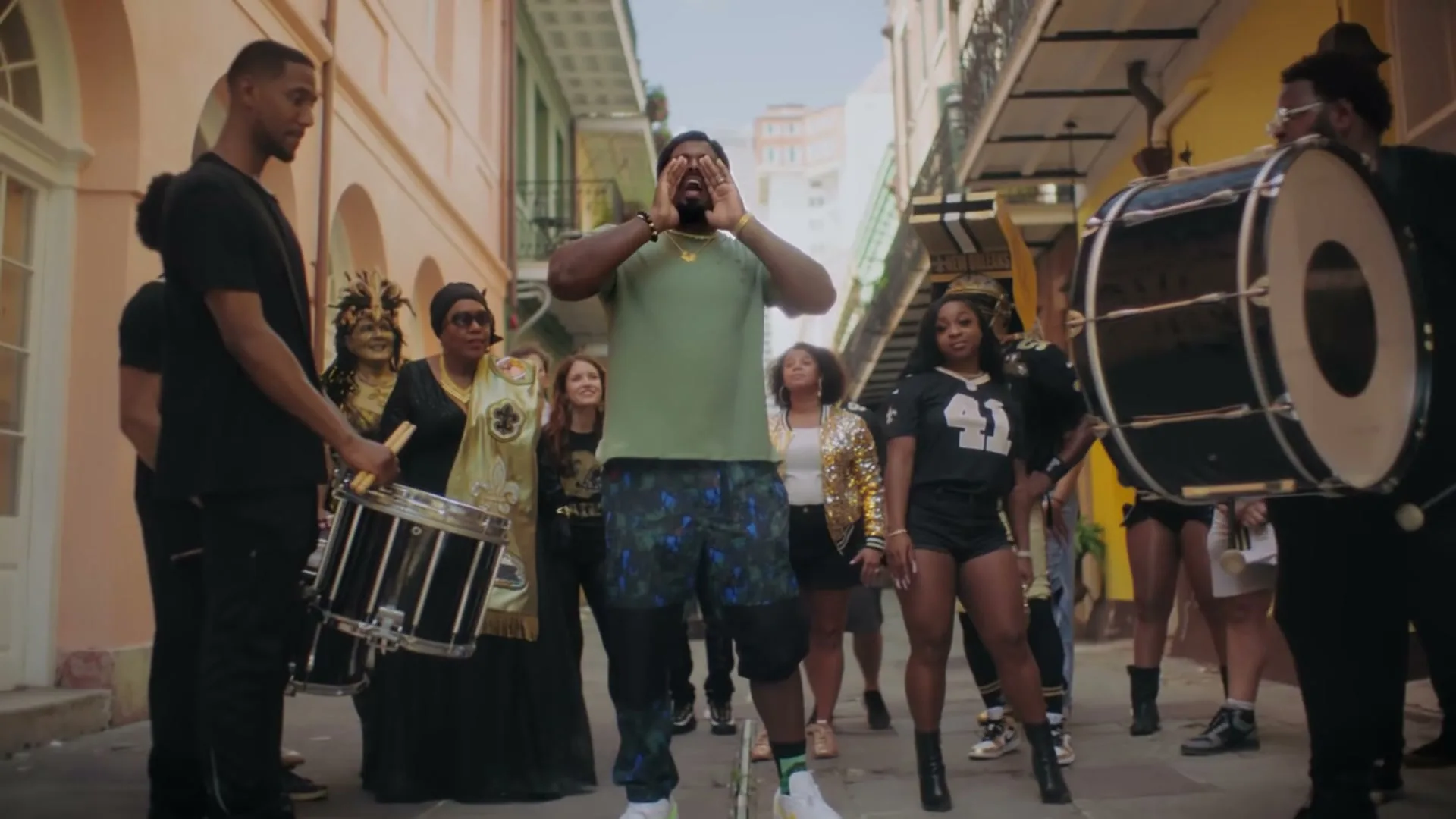 A man in a green shirt and patterned shorts shouts in a narrow street, surrounded by people in various outfits, including a marching band and a woman in a gold dress.
