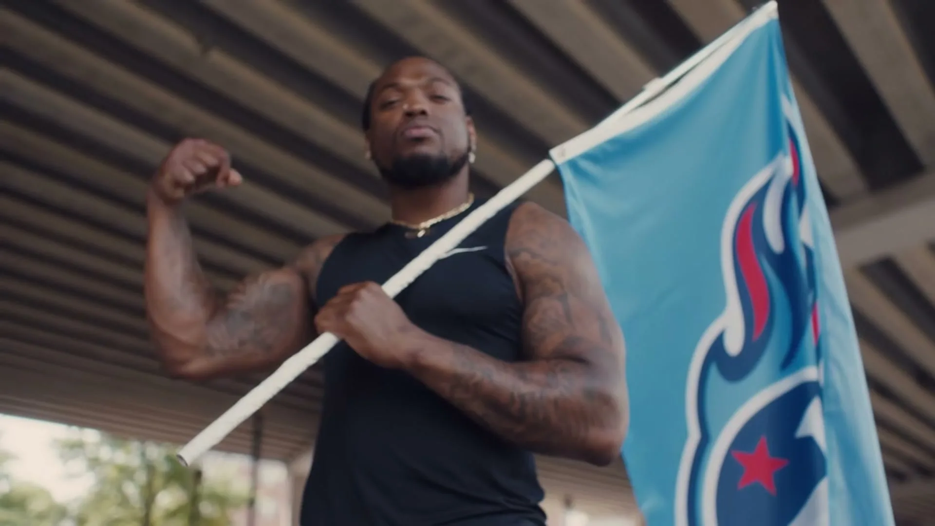 A muscular Black man in a black tank top holds a light blue flag with a red and white logo, flexing his arm under an overpass.