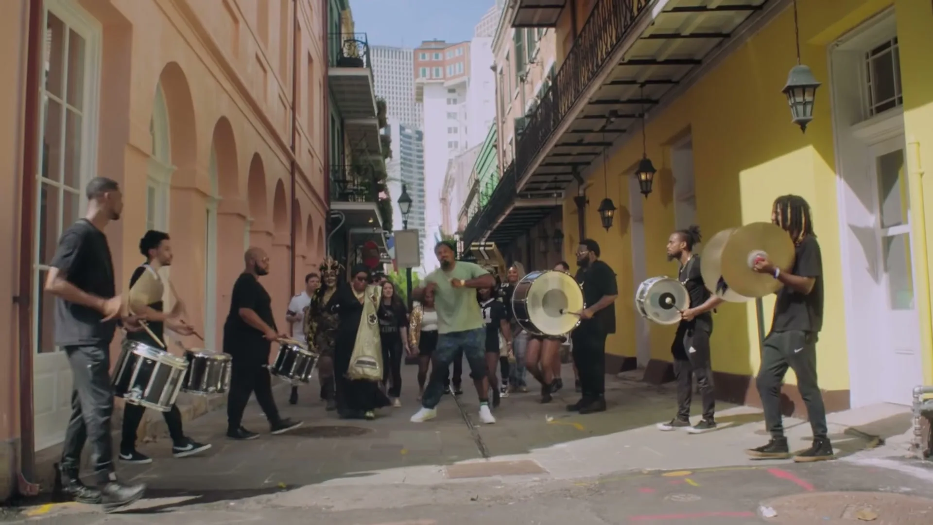 A group of musicians and dancers parade down a narrow street in a vibrant city, playing drums and cymbals.