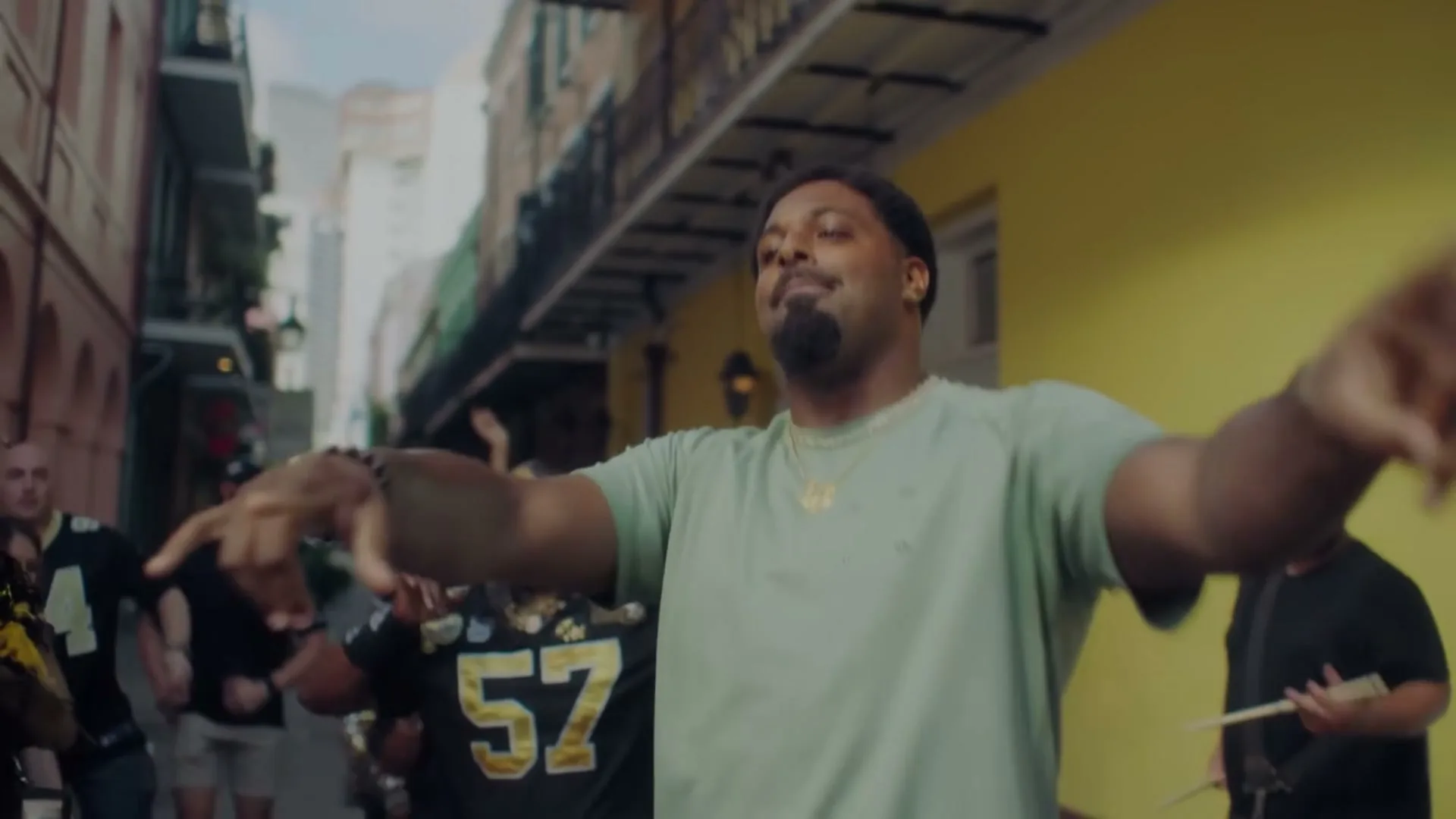 A man with a beard and a green shirt sings and dances in a street with a crowd of people in the background.