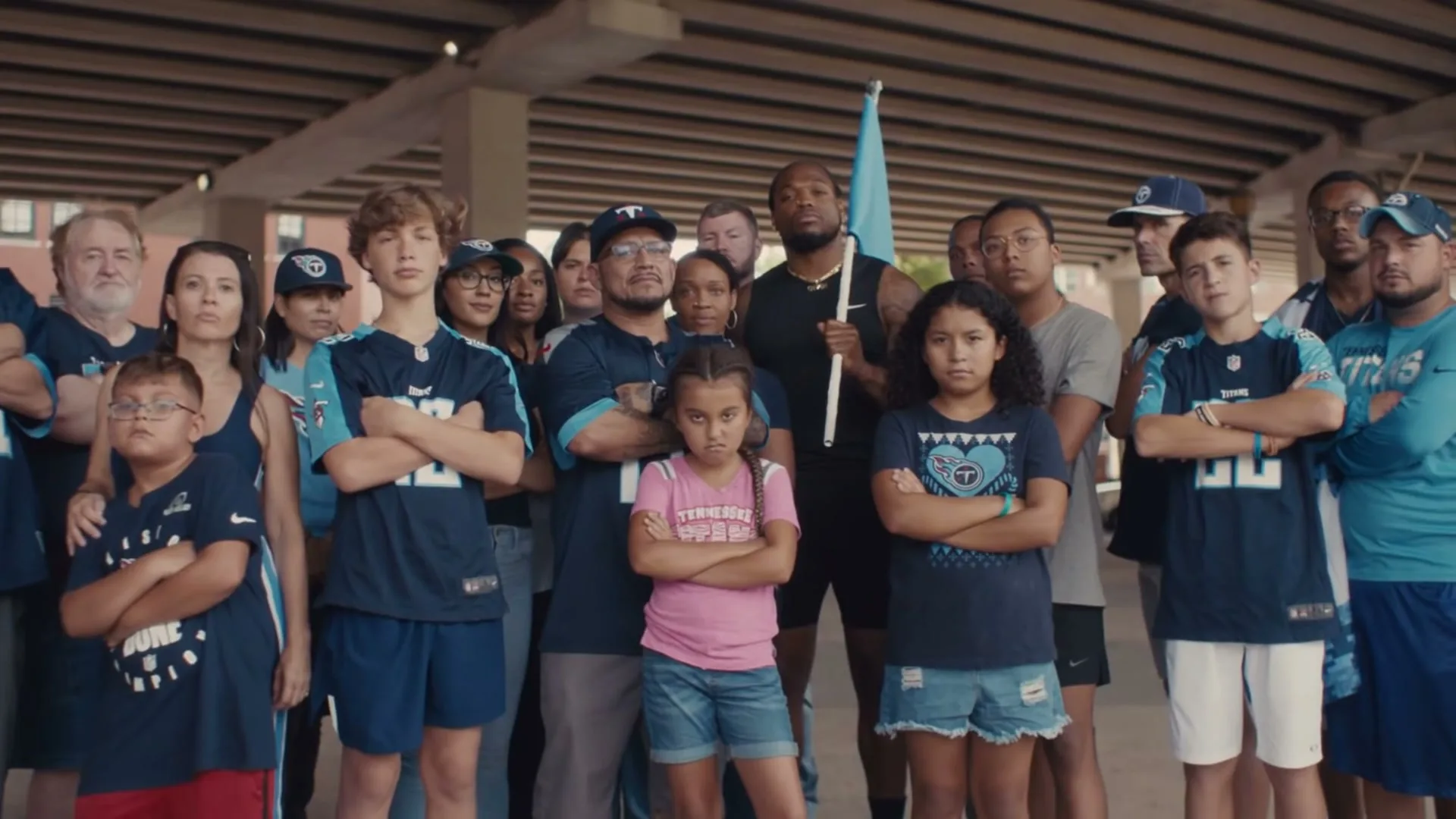 A diverse group of Tennessee Titans fans, including children and adults, stand with arms crossed under an overpass, looking directly at the camera.