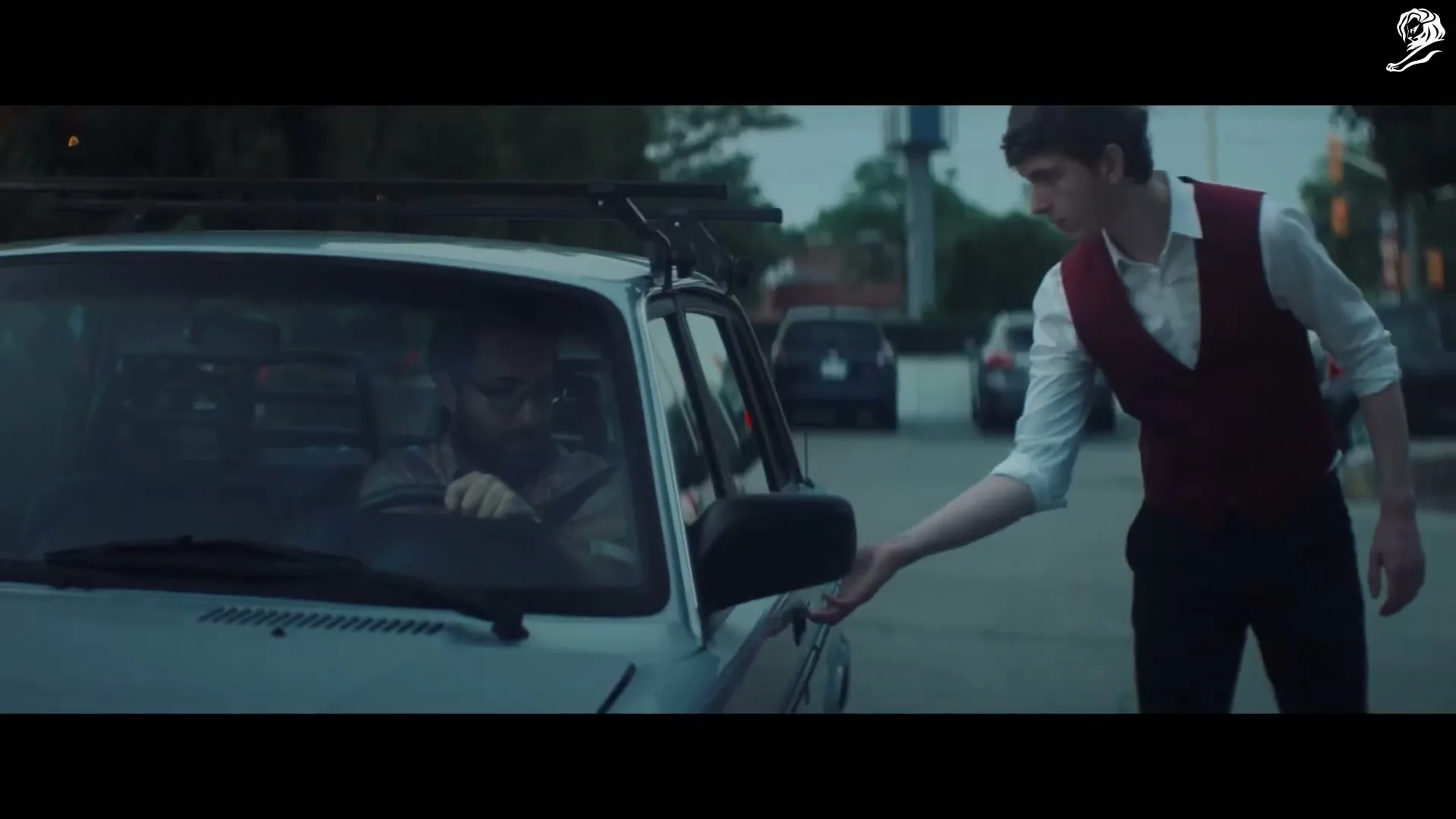 A young man in a white shirt and red vest reaches into a car to give something to a bearded man in glasses, who is sitting in the driver's seat.