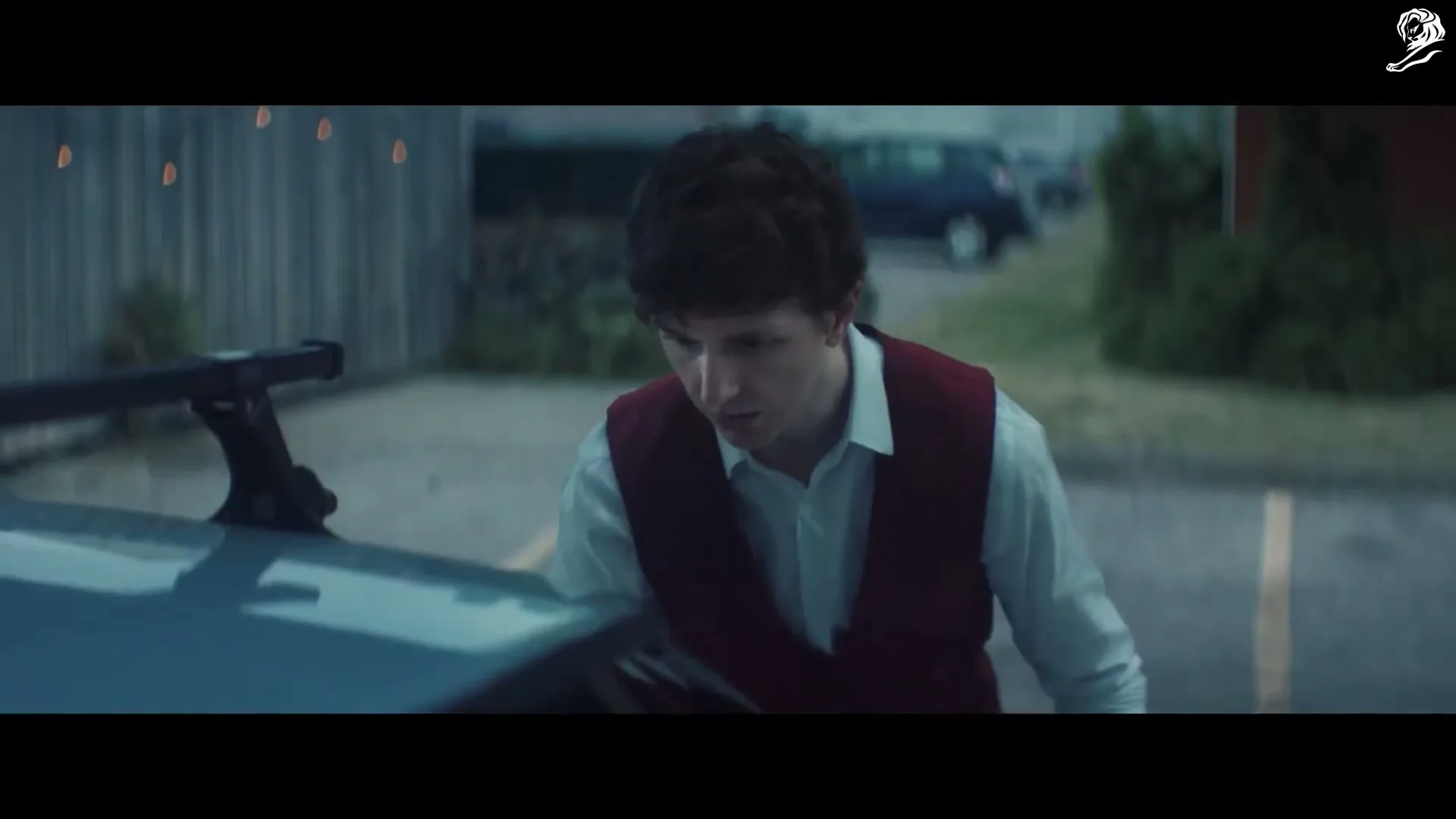 A young man with curly brown hair, wearing a white shirt and a red vest, looks down at the front of a car in a parking lot at dusk.