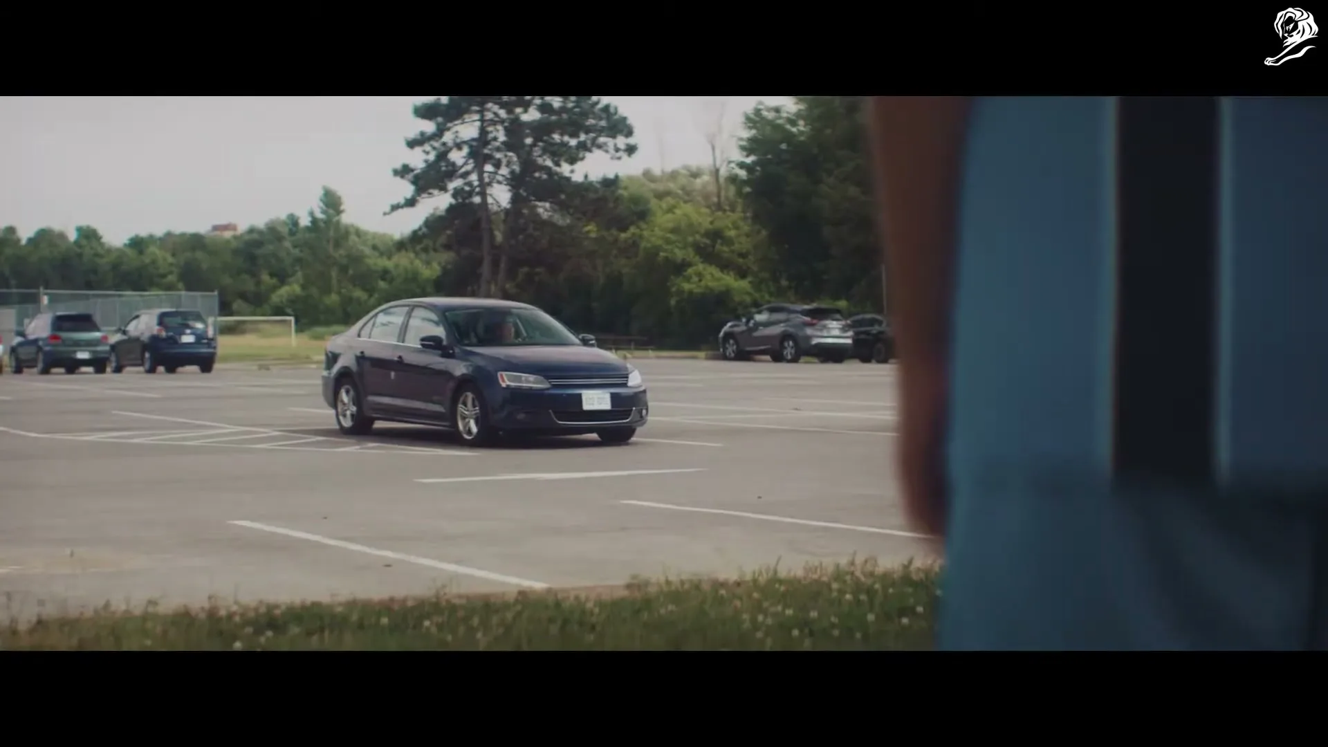 A dark blue sedan drives slowly through a mostly empty parking lot on a bright day, with a person's back visible in the foreground.