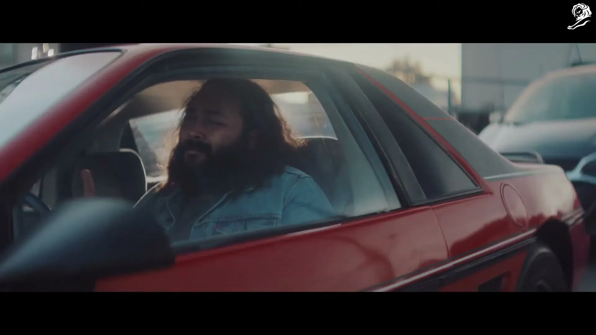A man with long hair and a beard, wearing a denim jacket, sits in a red car, singing along to music during the day.