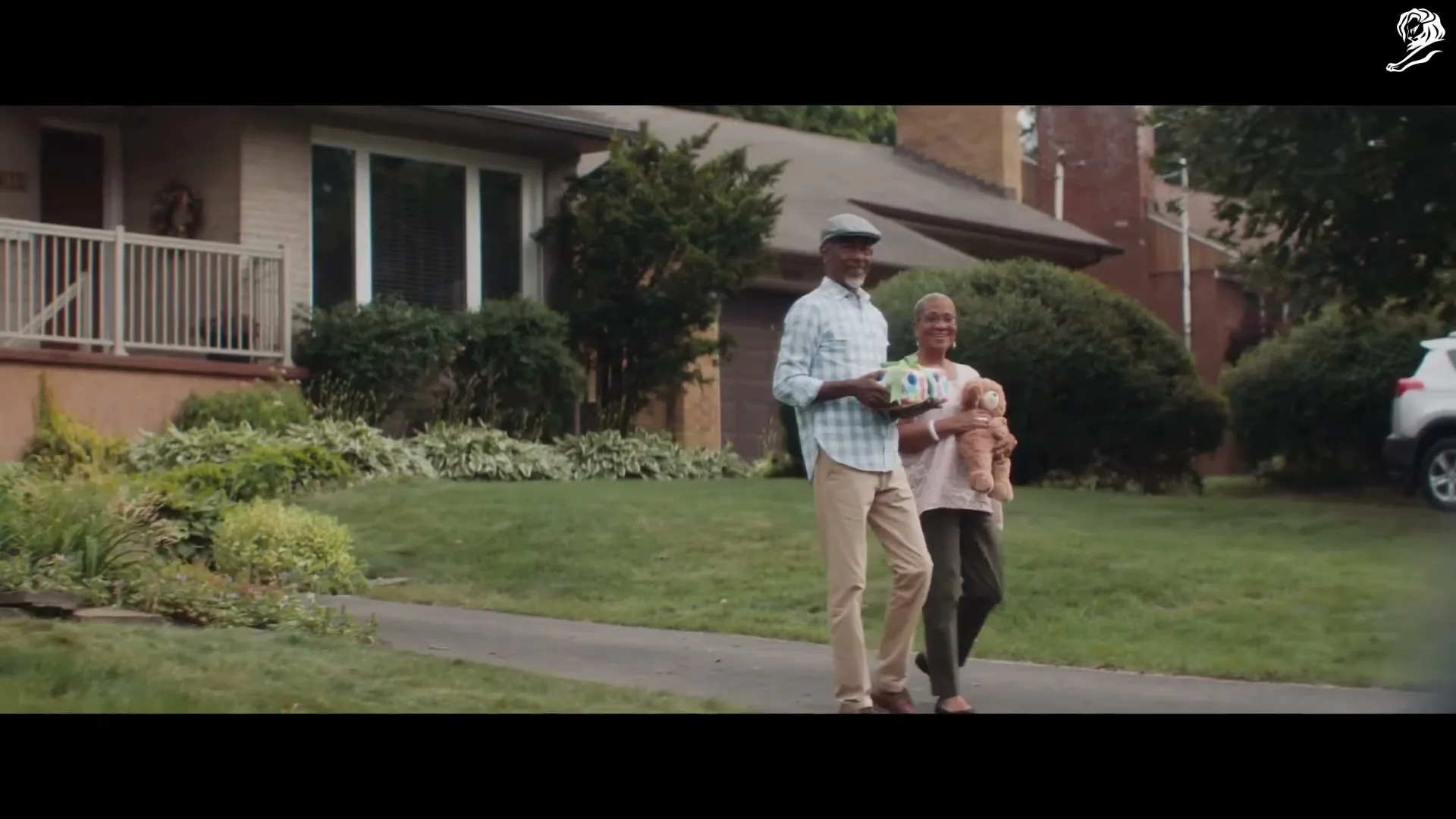 An older Black couple walks down a suburban driveway, the man holding a gift and the woman holding a doll.