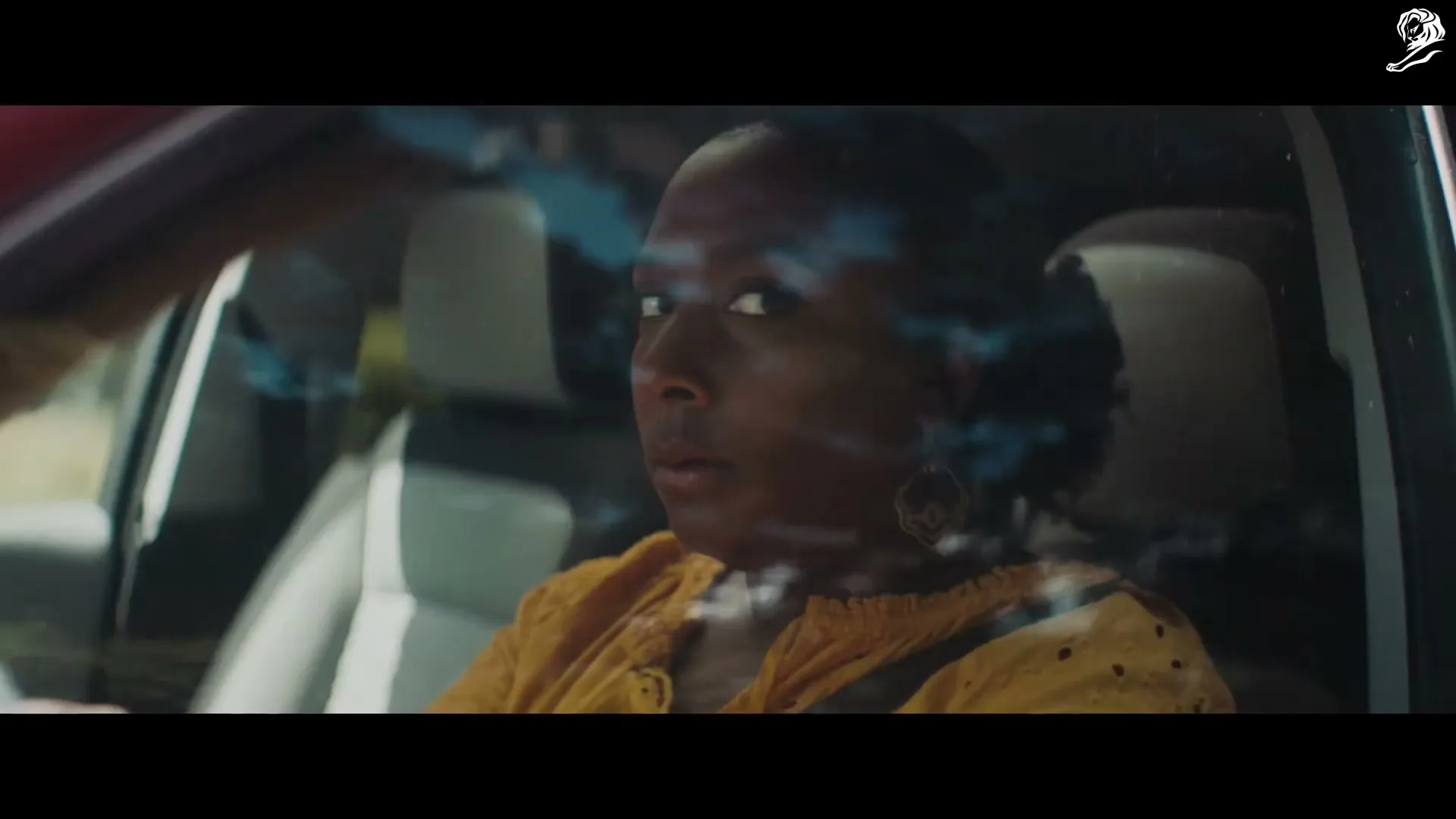 A Black woman in her 30s, wearing a yellow top, sits in the driver's seat of a car, looking out the window with a concerned expression.