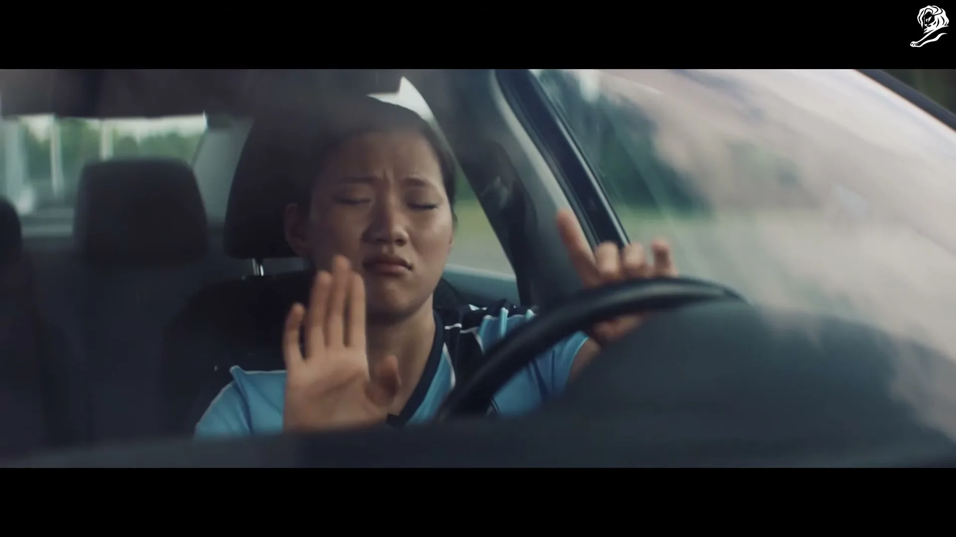 A young Asian woman in a blue shirt drives a car, looking distressed and holding her hand up as if to stop something.