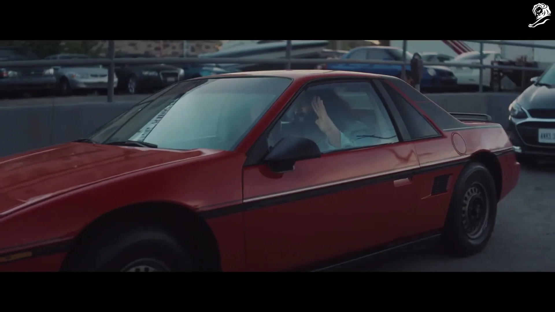A man with dark hair, wearing a light blue shirt, is driving a red sports car in a parking lot, looking around.