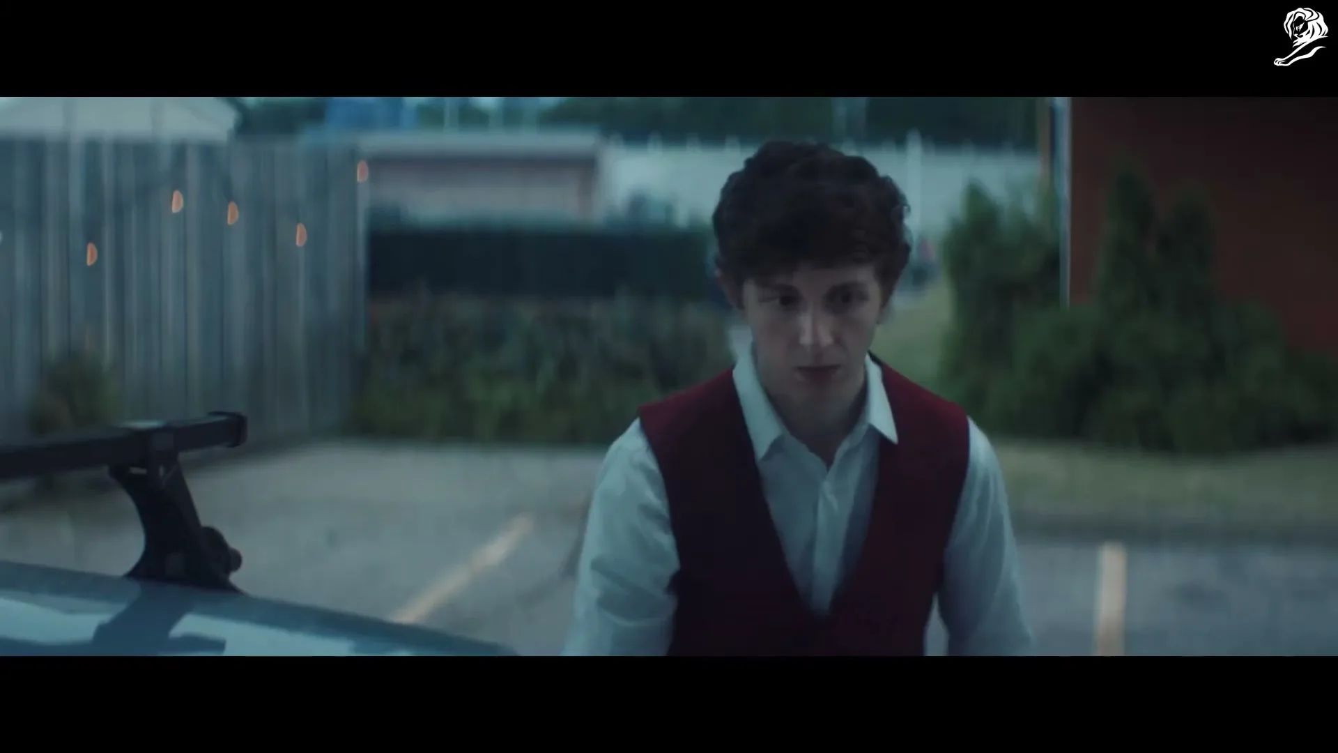 A young man with curly hair, wearing a white shirt and red vest, looks around nervously in a parking lot at dusk.