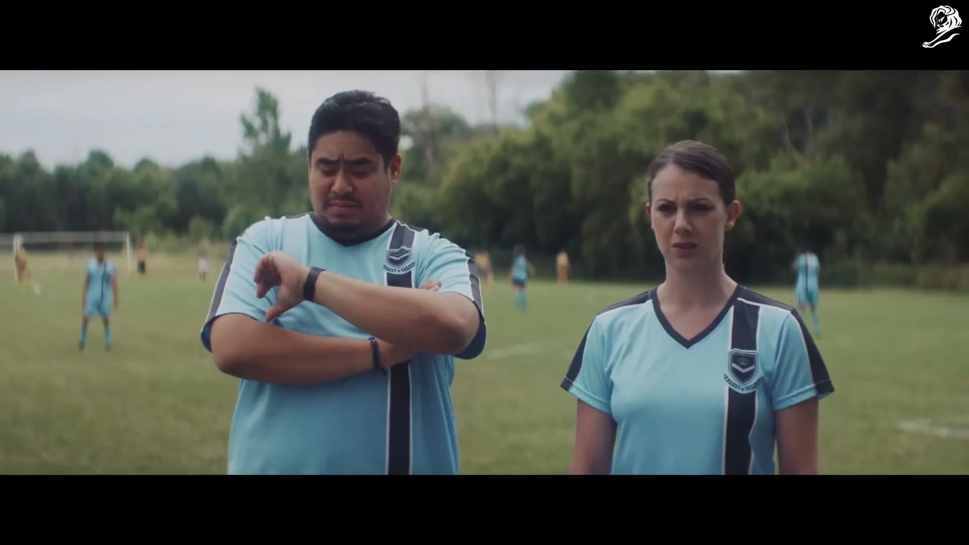 Two soccer coaches, a man and a woman, stand on a grassy field, looking confused as the man checks his watch.