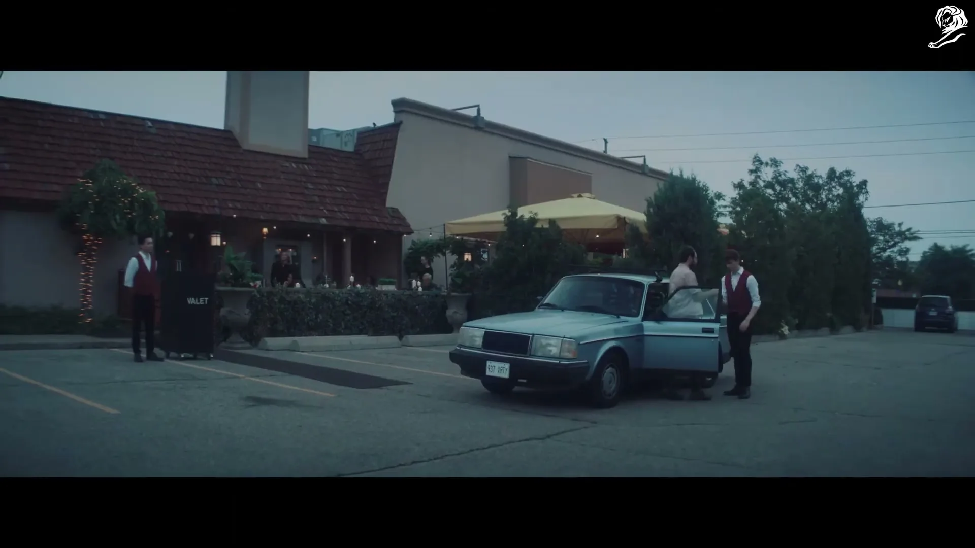 A man in a pink shirt and brown pants exits a light blue car in a parking lot, while two valets in red vests stand nearby outside a restaurant.