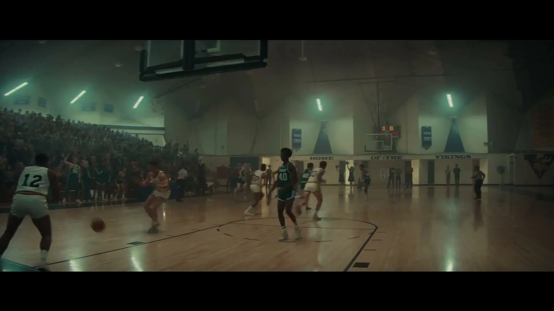 A wide shot of a basketball game in a high school gym, with players in green and white uniforms on the court and spectators in the stands.