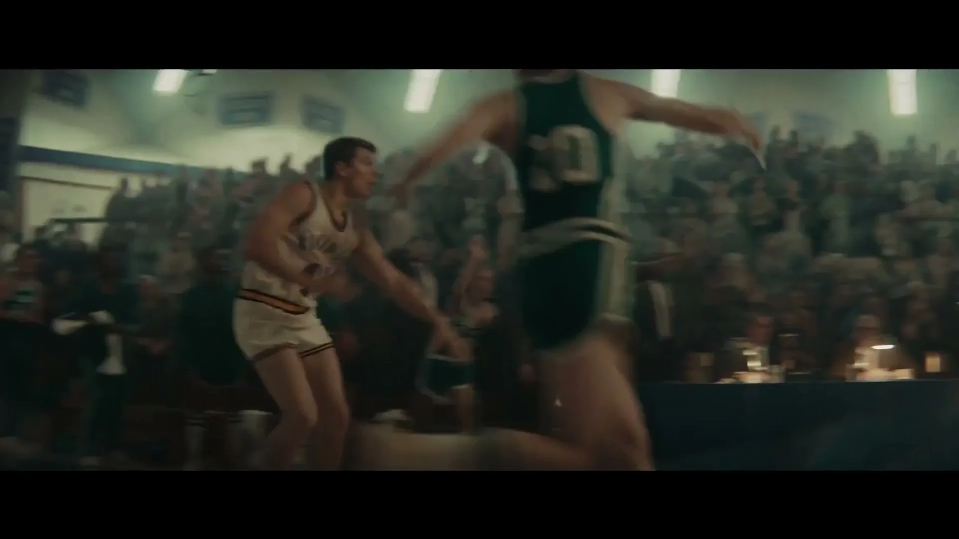 A basketball game is underway in a packed high school gym, with players in vintage uniforms dribbling and running across the court.