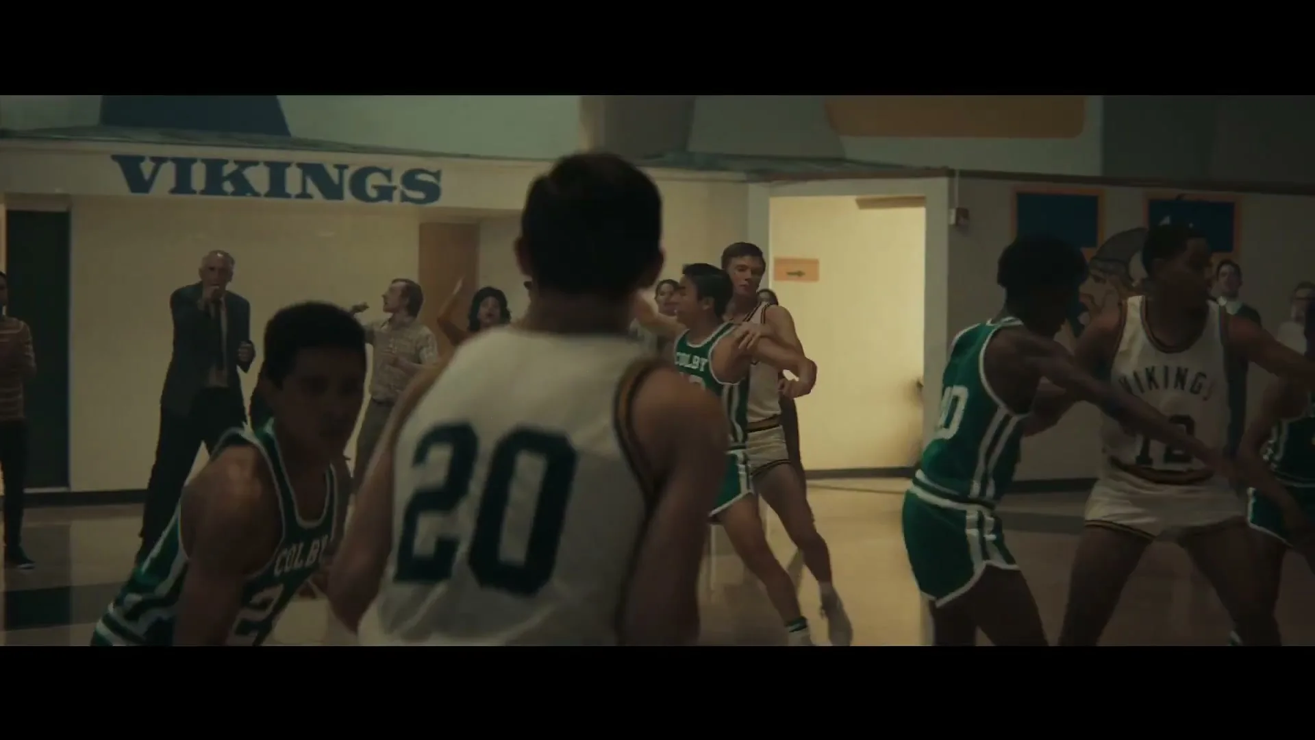 Basketball players in green and white jerseys play on a court while spectators watch from the sidelines in a high school gym.