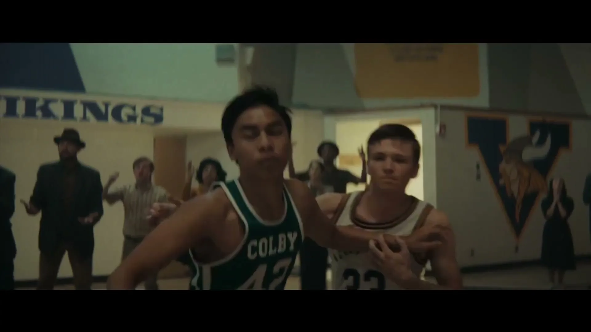 Two male basketball players, one in a green jersey and one in a white jersey, jostle for the ball on a basketball court while spectators watch in the background.