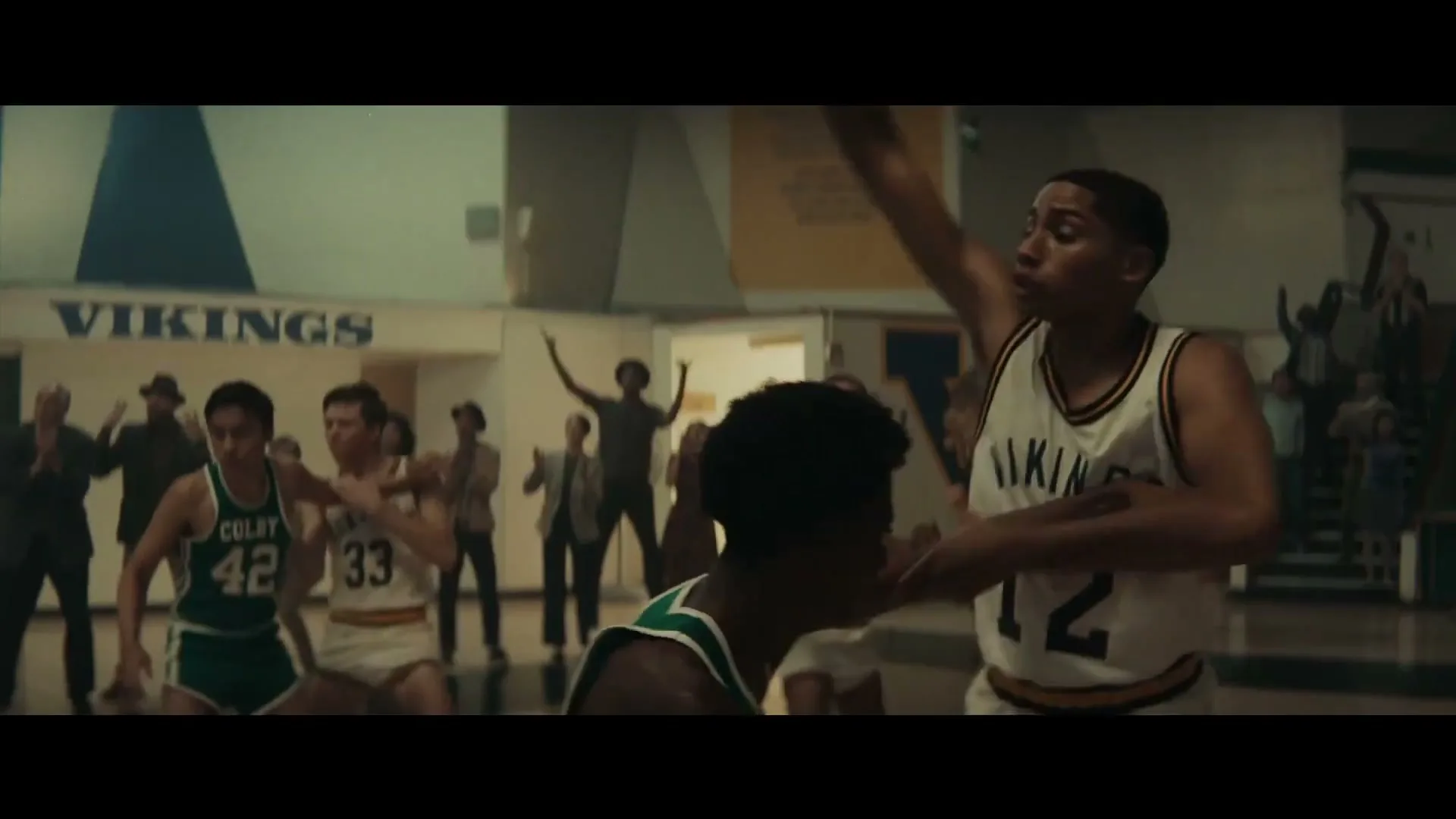 Two male basketball players in jerseys compete on a court while spectators watch in a high school gym.