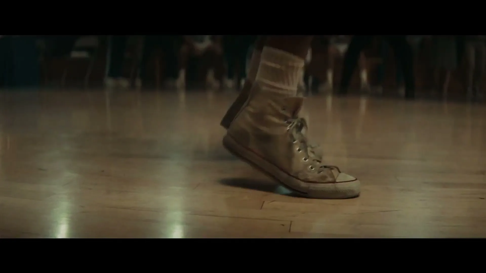 A person wearing white high-top sneakers and white socks jumps on a wooden basketball court, with blurred figures in the background.