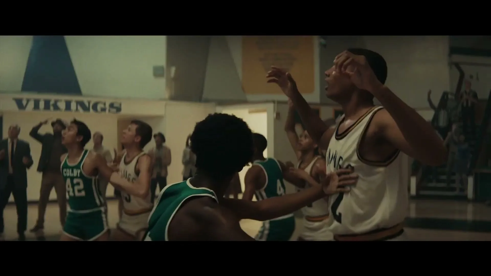 A group of young male basketball players in vintage uniforms look up intently during a game in a high school gym.