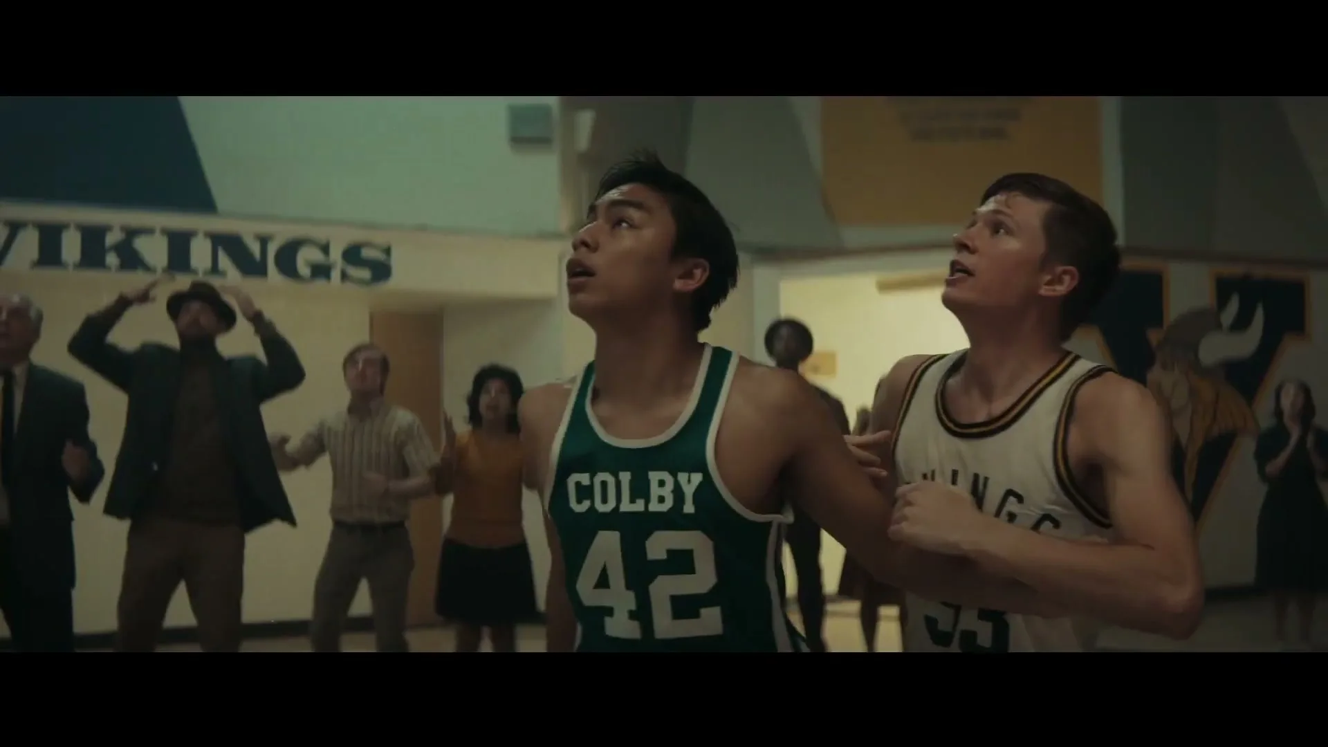 Two male basketball players, one in a green jersey and one in a white jersey, look up intently during a game in a high school gym.