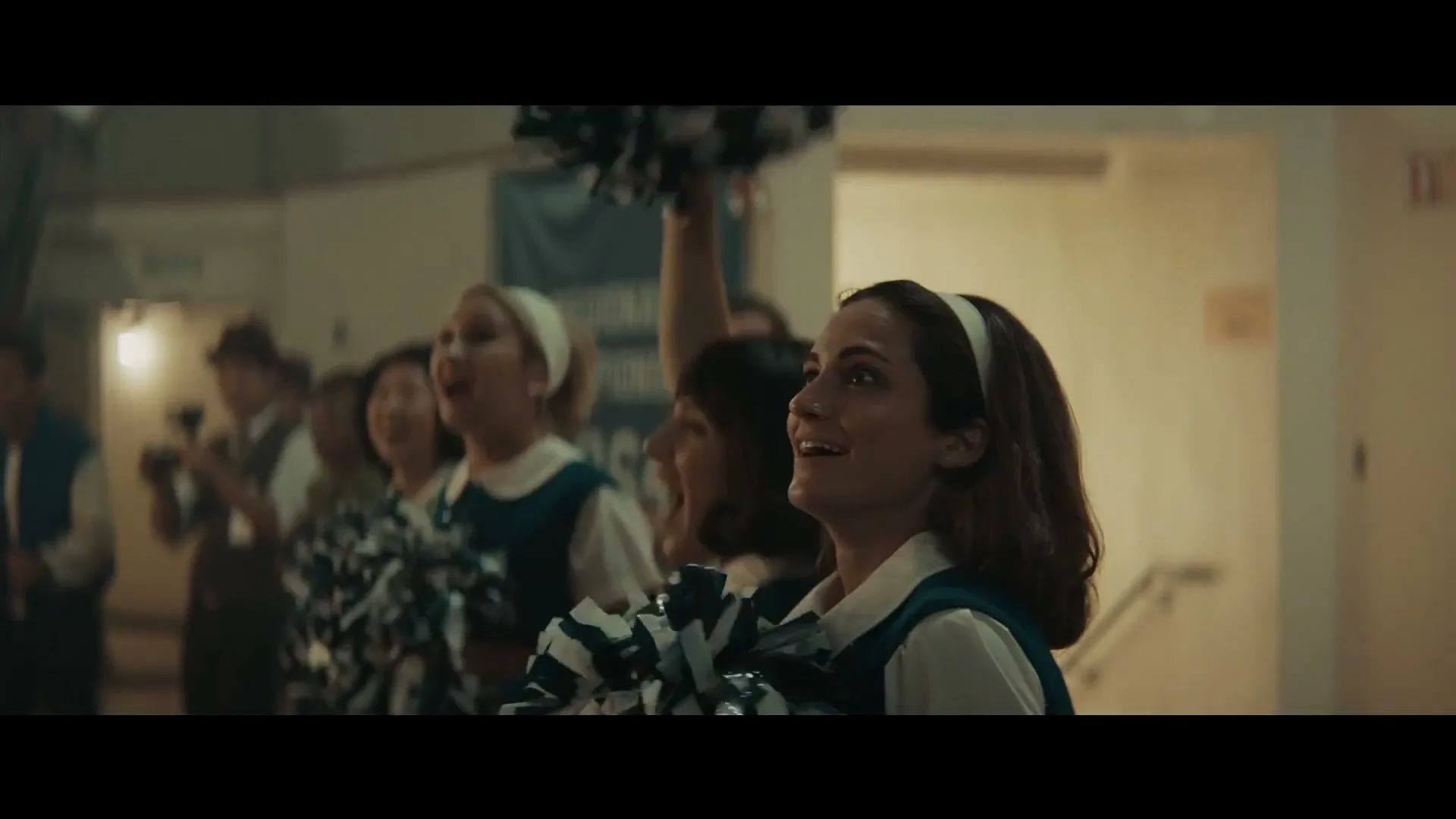 A group of female cheerleaders in vintage uniforms cheer enthusiastically, looking up and to the right, in a school hallway.
