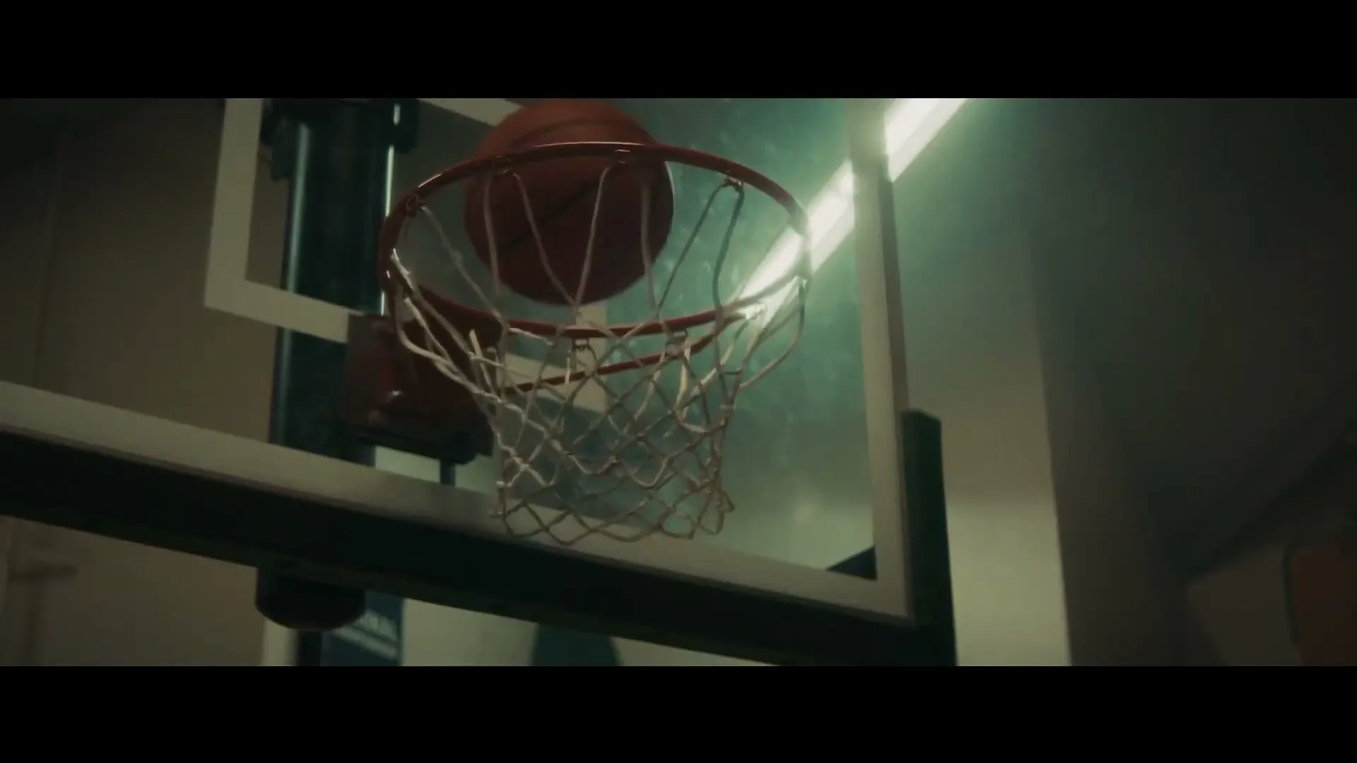 A basketball swishes through a hoop, with bright light shining from behind the backboard in a dimly lit gym.