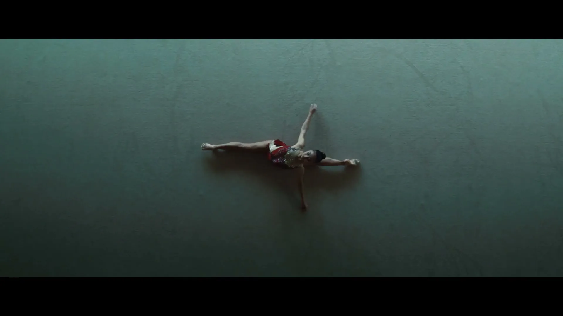 A young female gymnast in a sparkly leotard lies on a light green mat, stretching her limbs out, viewed from directly above.